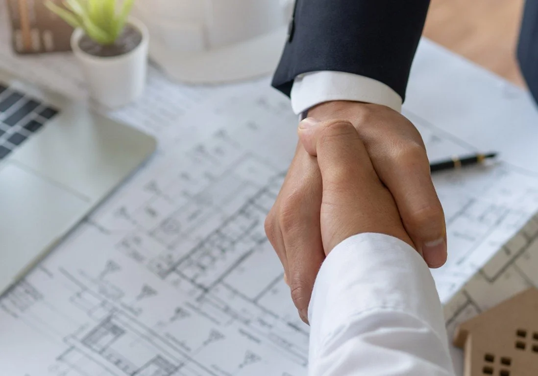 Close-up of two people shaking hands over a desk with architectural blueprints, a pen, a laptop, and a small plant.