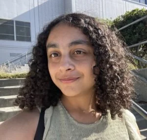 A young woman with curly hair smiling slightly outdoors near a modern glass building and stairs.