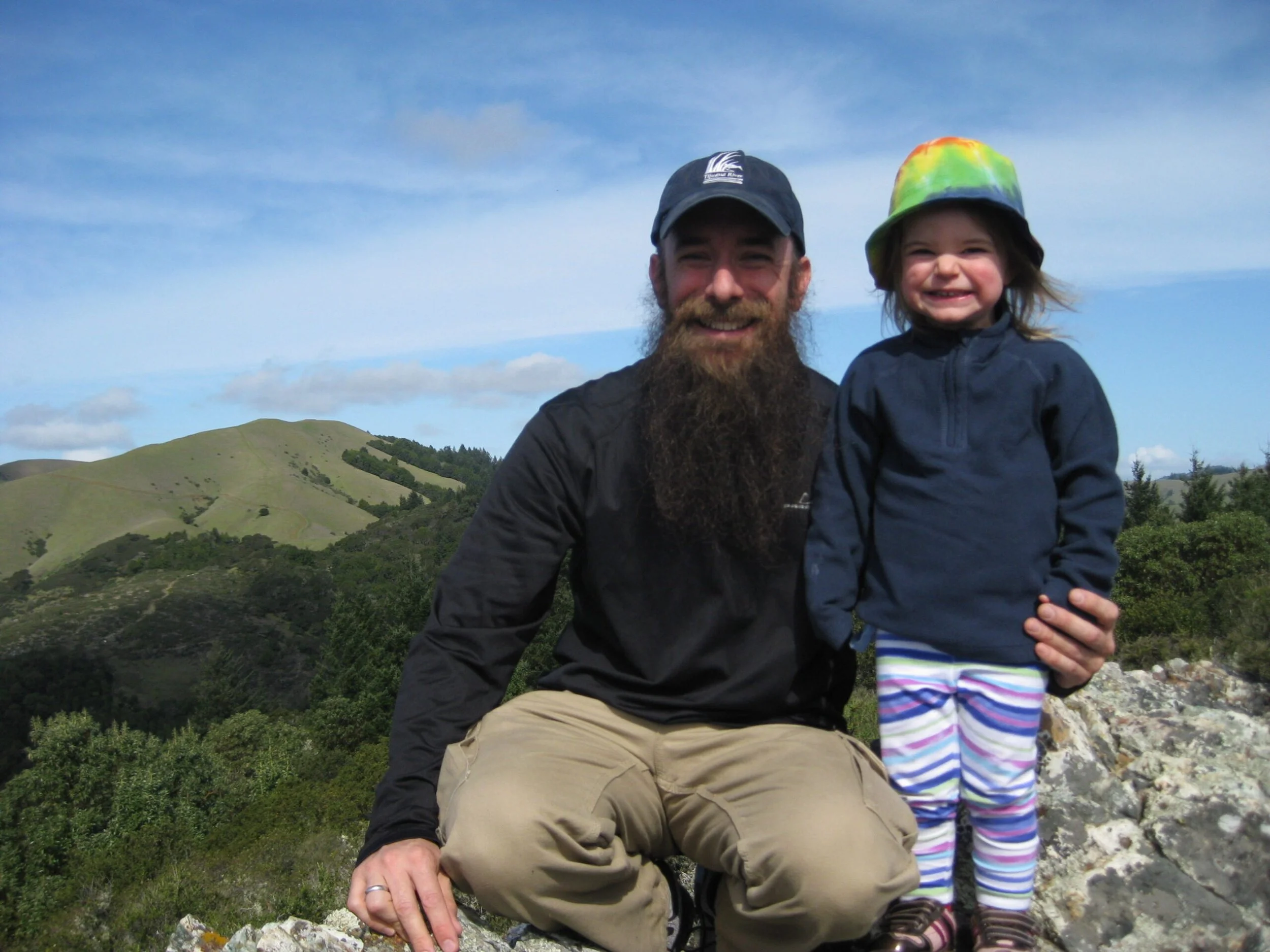 A man with a beard and a child wearing a colorful tie-dye hat and striped leggings, both smiling, outdoors with green rolling hills and a blue sky with clouds in the background.