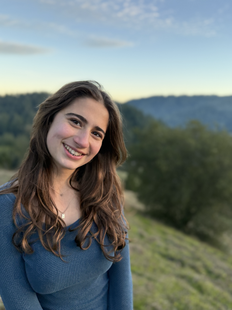 Young woman with long brown hair smiling outdoors, wearing a blue sweater, standing in a scenic natural landscape with trees and hills in the background during daytime.