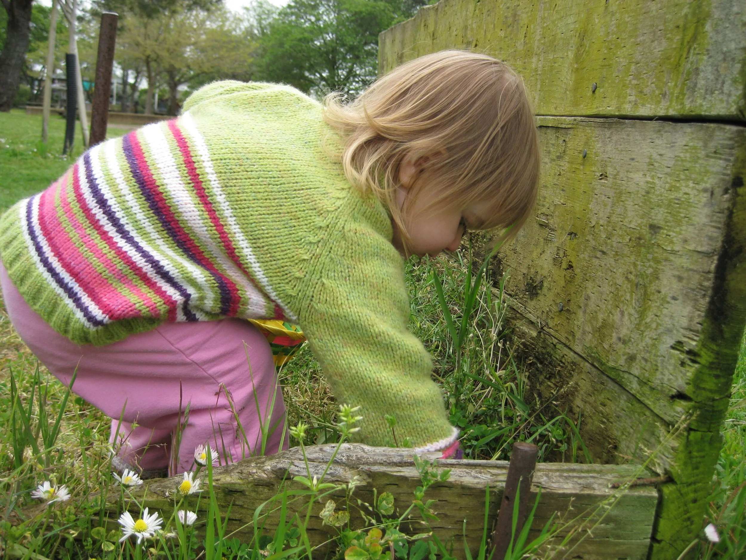 A young girl with reddish hair in a braided ponytail, wearing a green sweater with colorful stripes and pink pants, crouching outdoors near a moss-covered wooden fence.