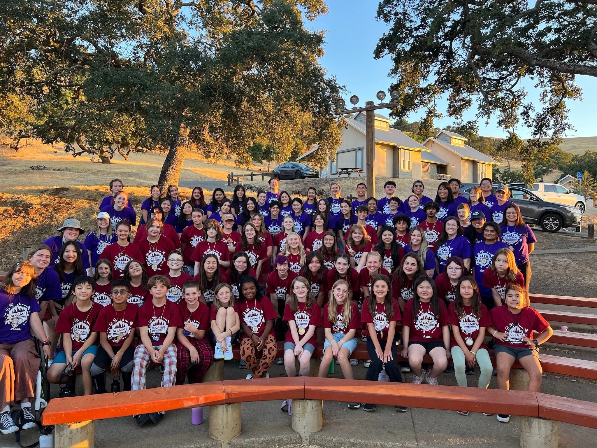 A large group of children and teenagers gathered outdoors for a photo during a camp, with some wearing matching purple and red T-shirts that say 'JA Camps.' The children are smiling and standing in front of a backdrop of trees, rustic buildings, and parked cars, with the sun casting warm light.