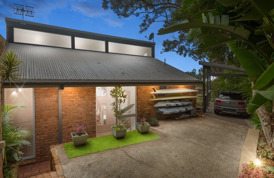 A house with a brick exterior and a sloped corrugated metal roof, surrounded by tropical plants, with a driveway holding a carport with a sports car and a black car, and a storage rack with kayaks or paddleboards.