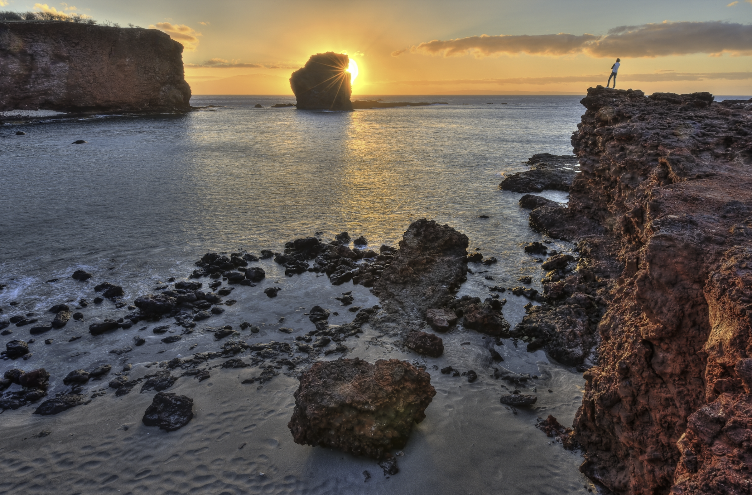 A scenic ocean view at sunset with rocky cliffs and a person standing on the edge of one cliff.