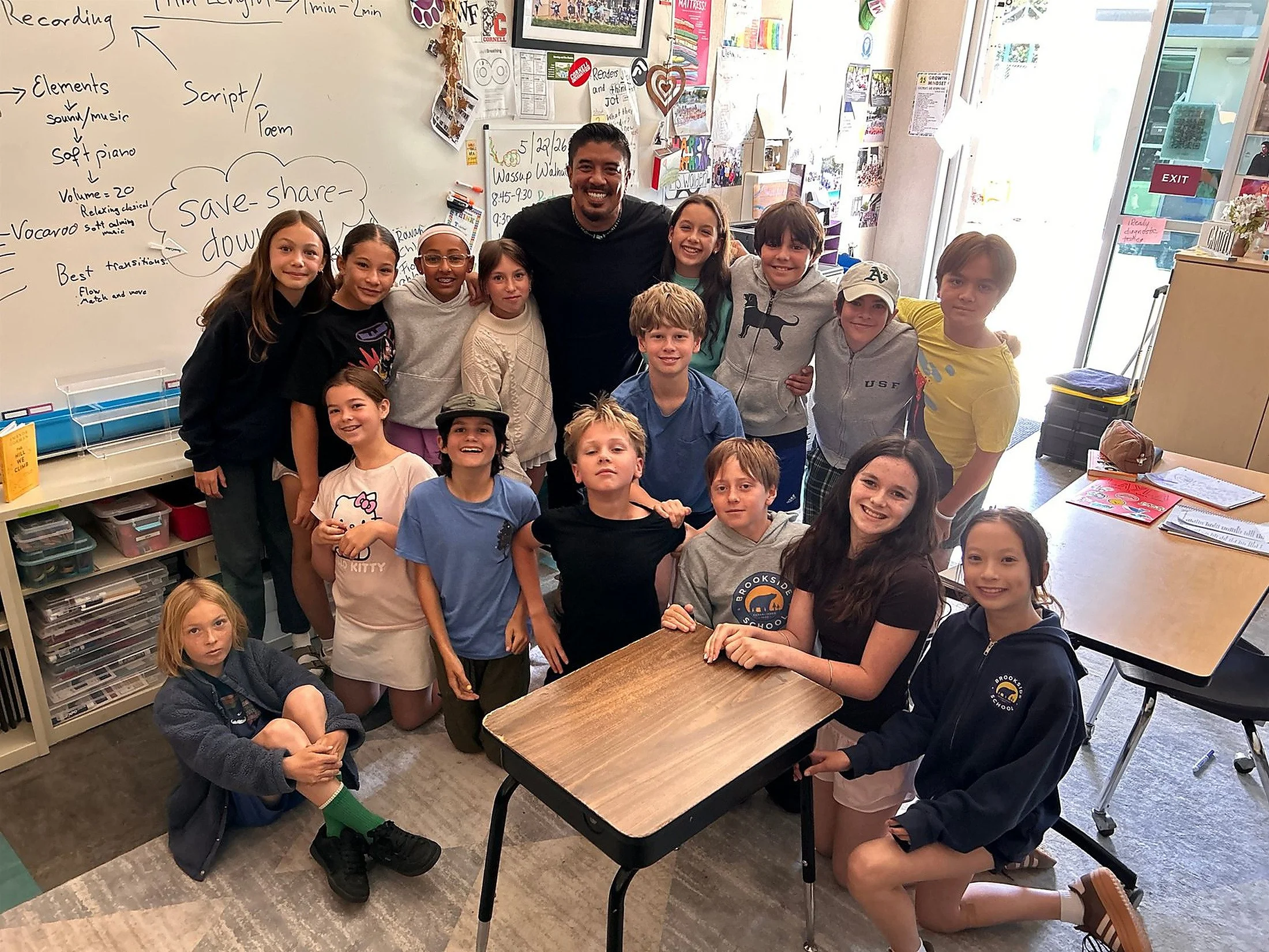 A group of children and a man are smiling and posing for a photo in a classroom. The classroom has a whiteboard with notes and drawings, and there are posters, artwork, and a door with windows in the background.