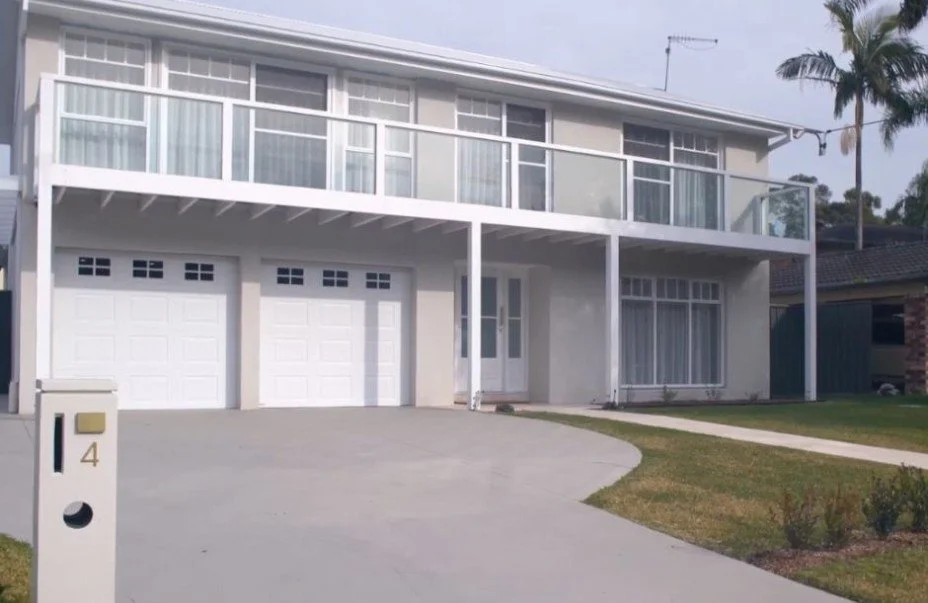 Modern two-story house with a white exterior, glass balcony railing, and a three-car garage. There is a mailbox with the number 4 and a lawn with a concrete pathway in front.