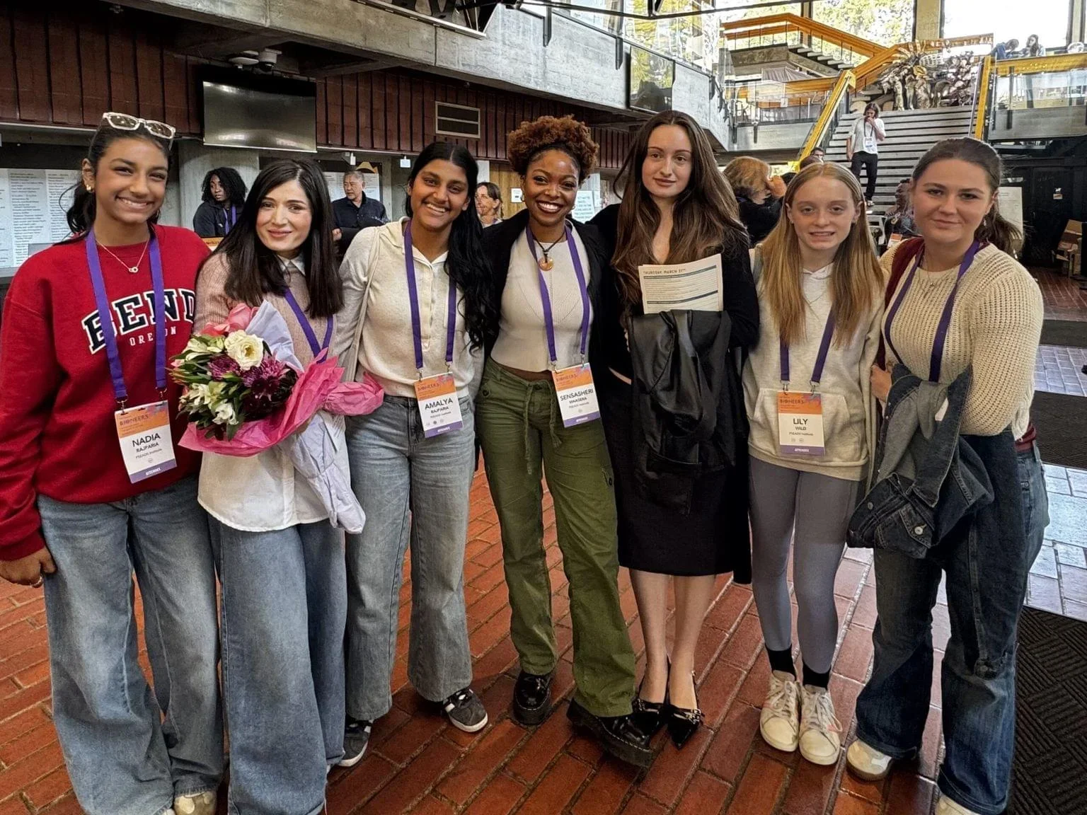 Group of seven diverse young women standing together indoors, smiling, some holding flowers and papers, wearing conference badges on lanyards.