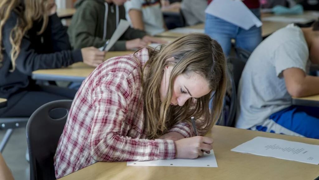 A young woman with long brown hair wearing a red checked shirt is sitting at a desk, writing on a sheet of paper during a classroom exam.