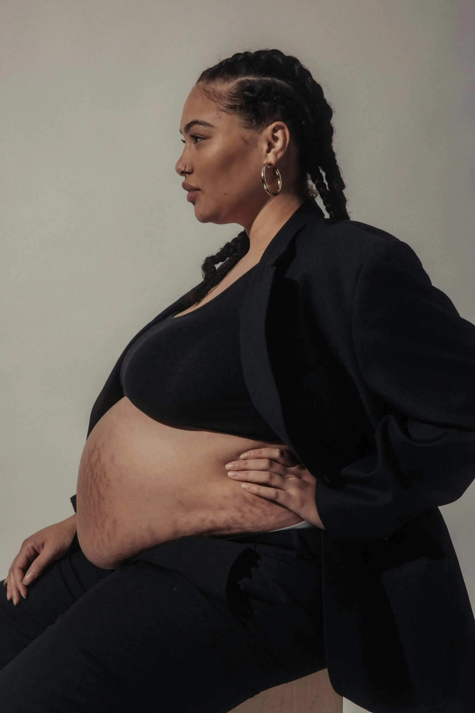 Side profile of a woman with styled braids, wearing hoop earrings, a black blazer and top, and black pants, sitting with one hand on her hip against a plain background.