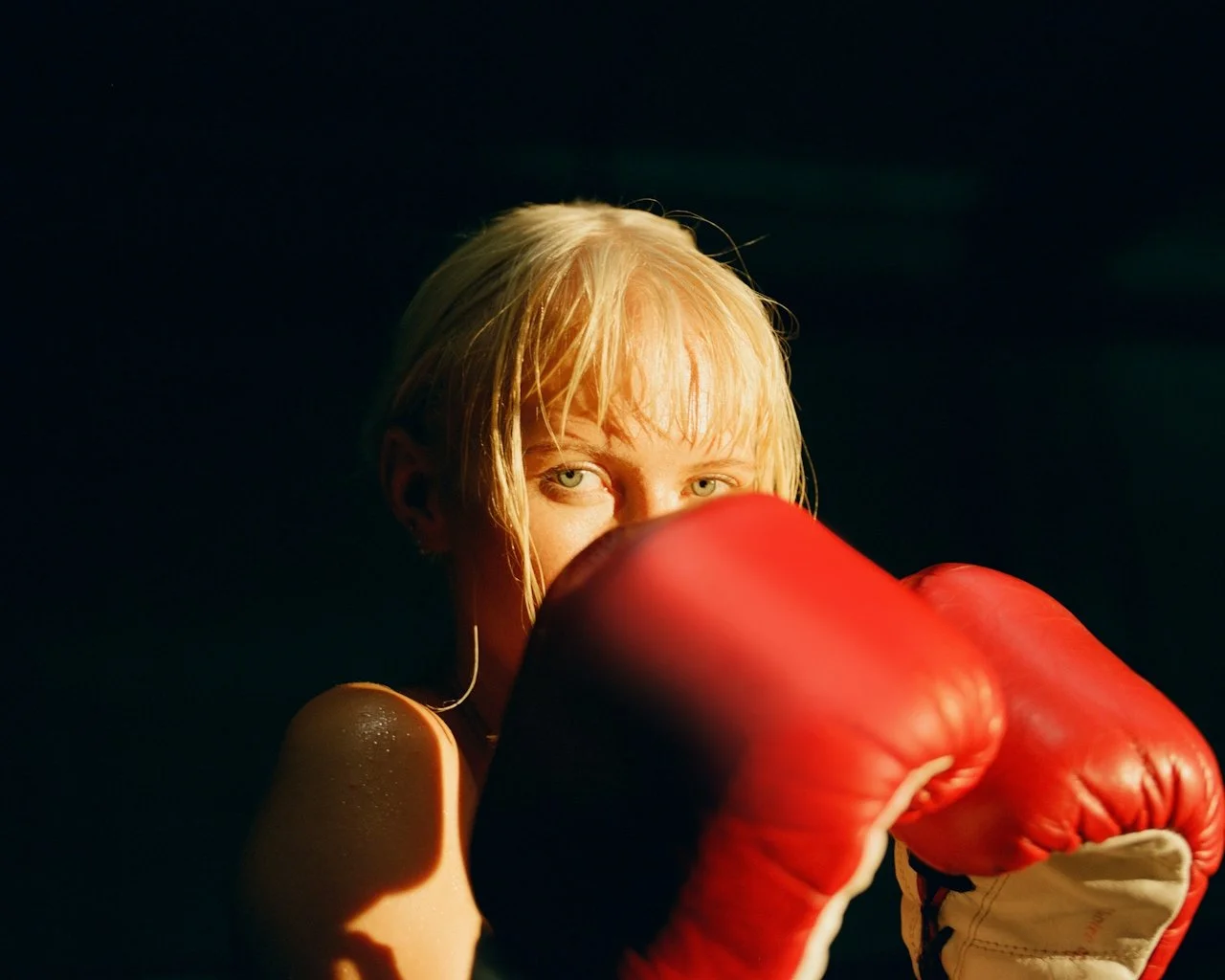 A woman with blonde hair and light-colored eyes wearing red boxing gloves, partially obscuring her face, in a dark setting with dramatic lighting.