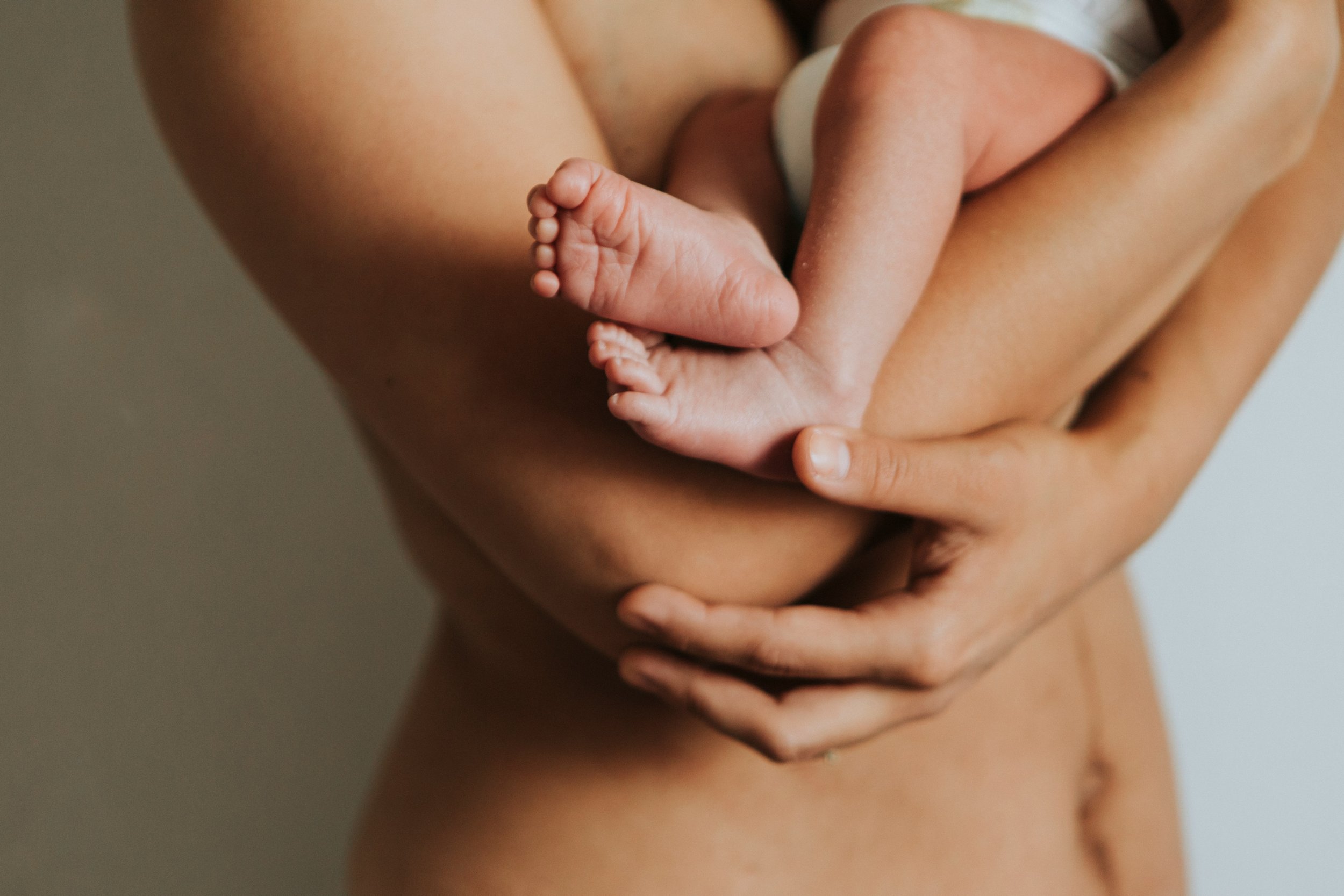 Close-up of an adult holding a newborn baby against their chest, with focus on the baby's tiny feet and the adult's arms.