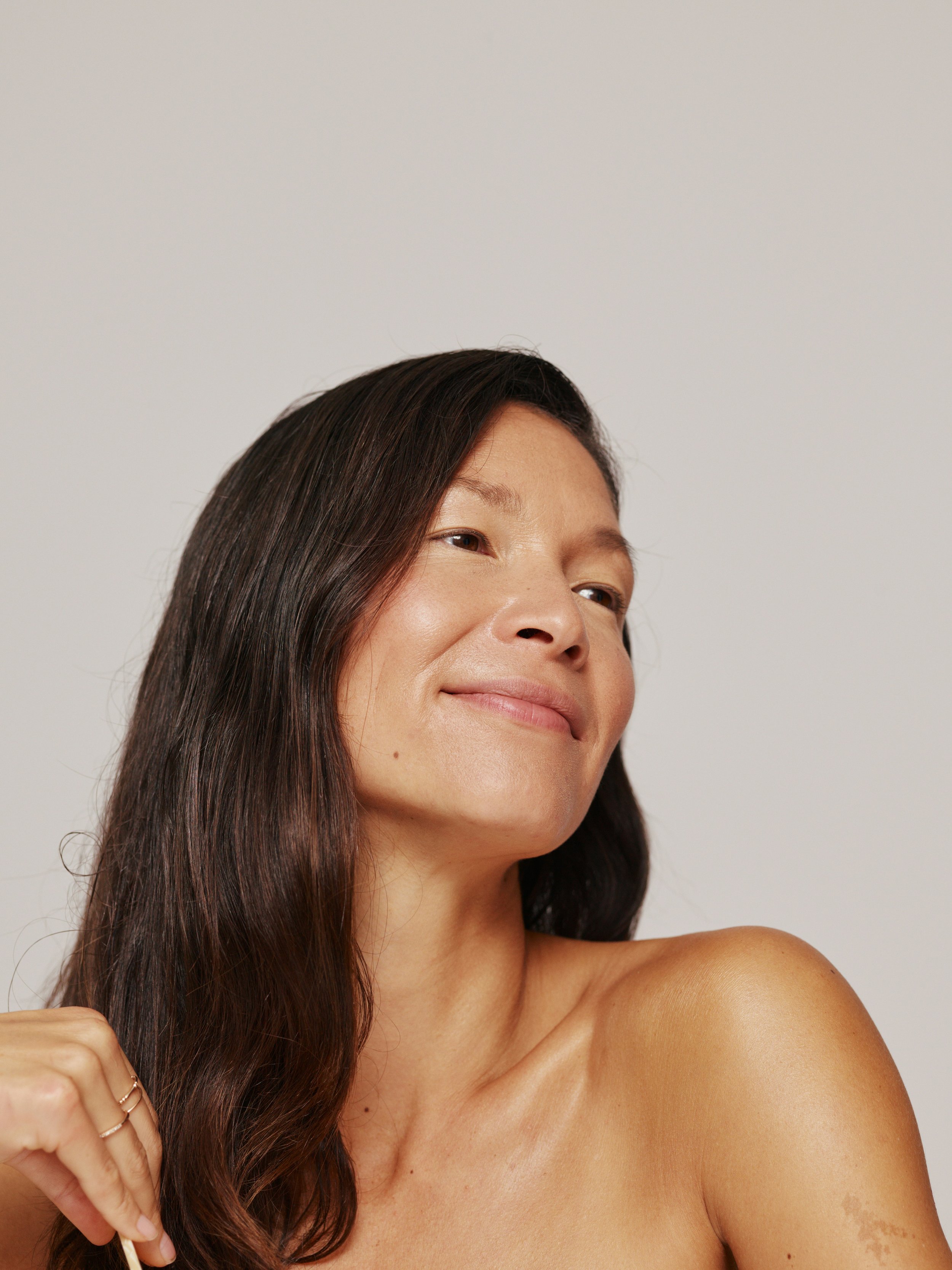 A woman with long, wavy brown hair smiling gently against a plain, off-white background.