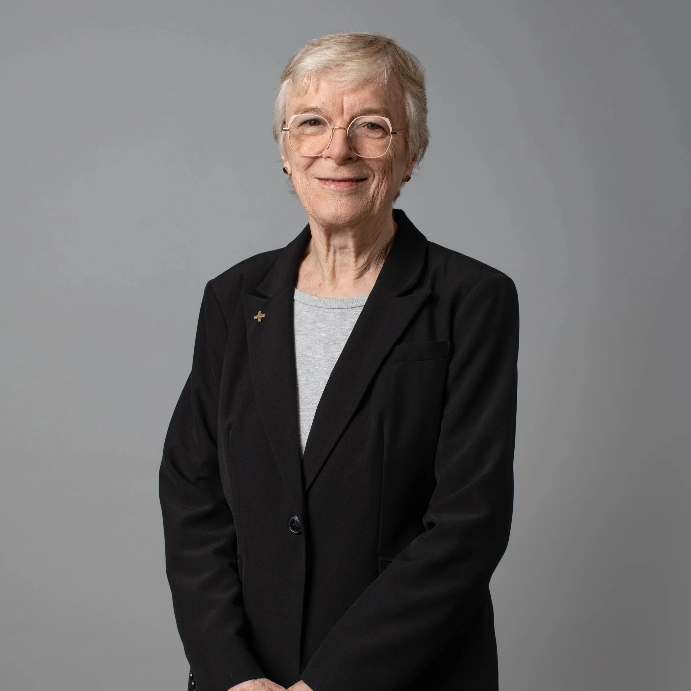 An elderly woman with short gray hair, wearing glasses, a black blazer, and a light gray shirt, standing against a blue background.