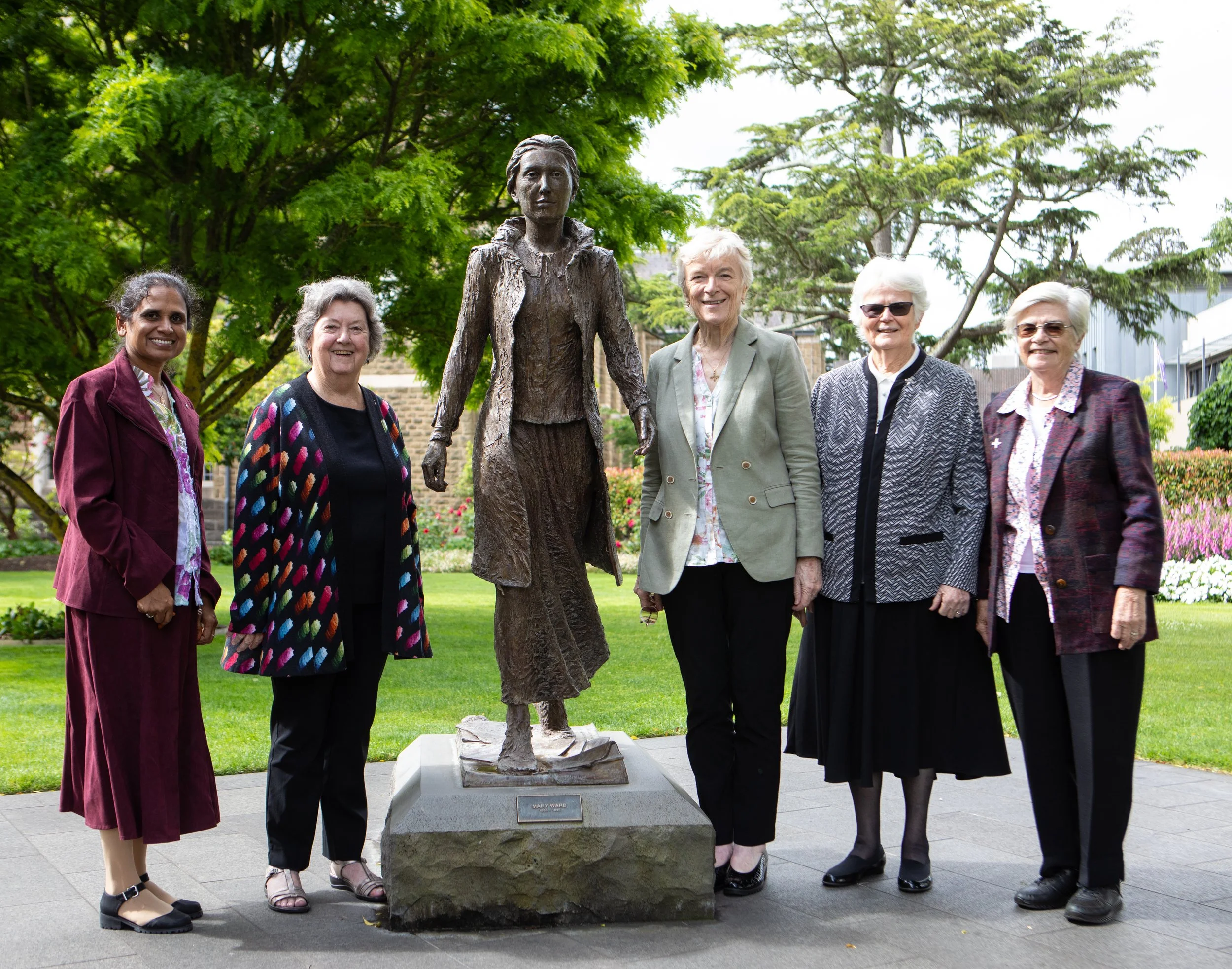 Six women standing outdoors in a park, posing around a bronze statue of a woman. The women are smiling and dressed in various colorful and neutral outfits. The background features green trees and a manicured garden.