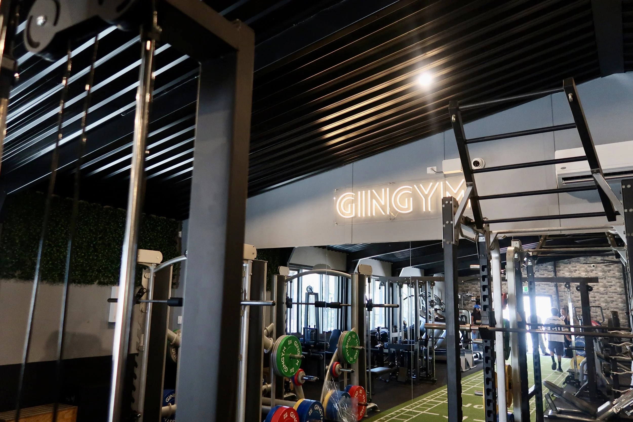 Interior of a gym with workout equipment, including weight racks with colorful weights, a pull-up bar, and a mirror reflecting the gym space. The ceiling has black slats with a bright light and a neon sign that reads 'GINGY'.