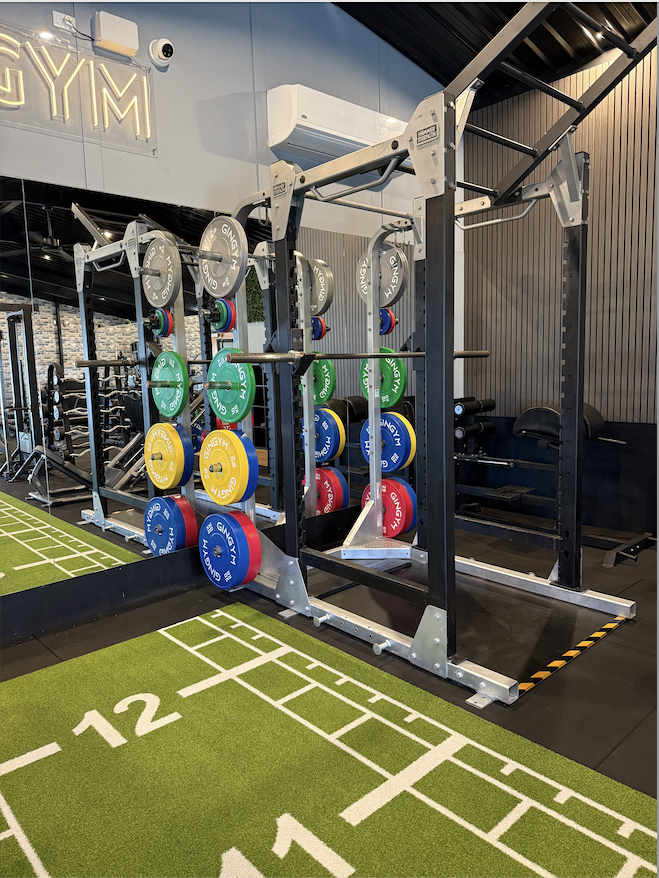 A gym with a power rack, colorful weight plates, and a green exercise mat with white lines and numbers.