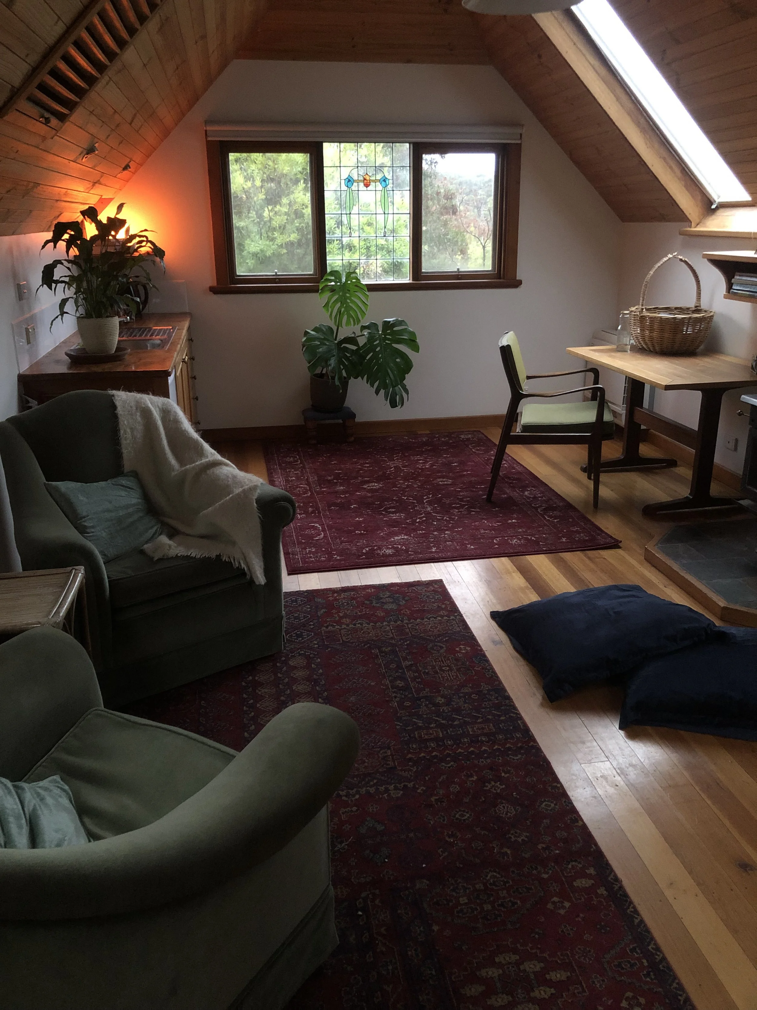 A cozy attic room with wooden vaulted ceiling, window with stained glass, green armchair, sofa with white blanket, red patterned rug, indoor plants, and skylight.