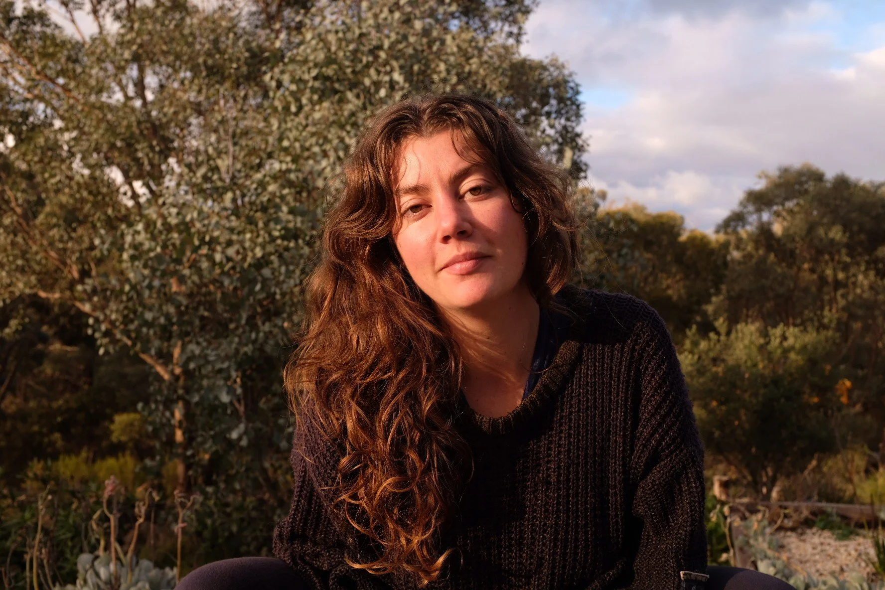 A woman with long curly hair sitting outdoors during late afternoon or sunset, with trees and a partly cloudy sky in the background.