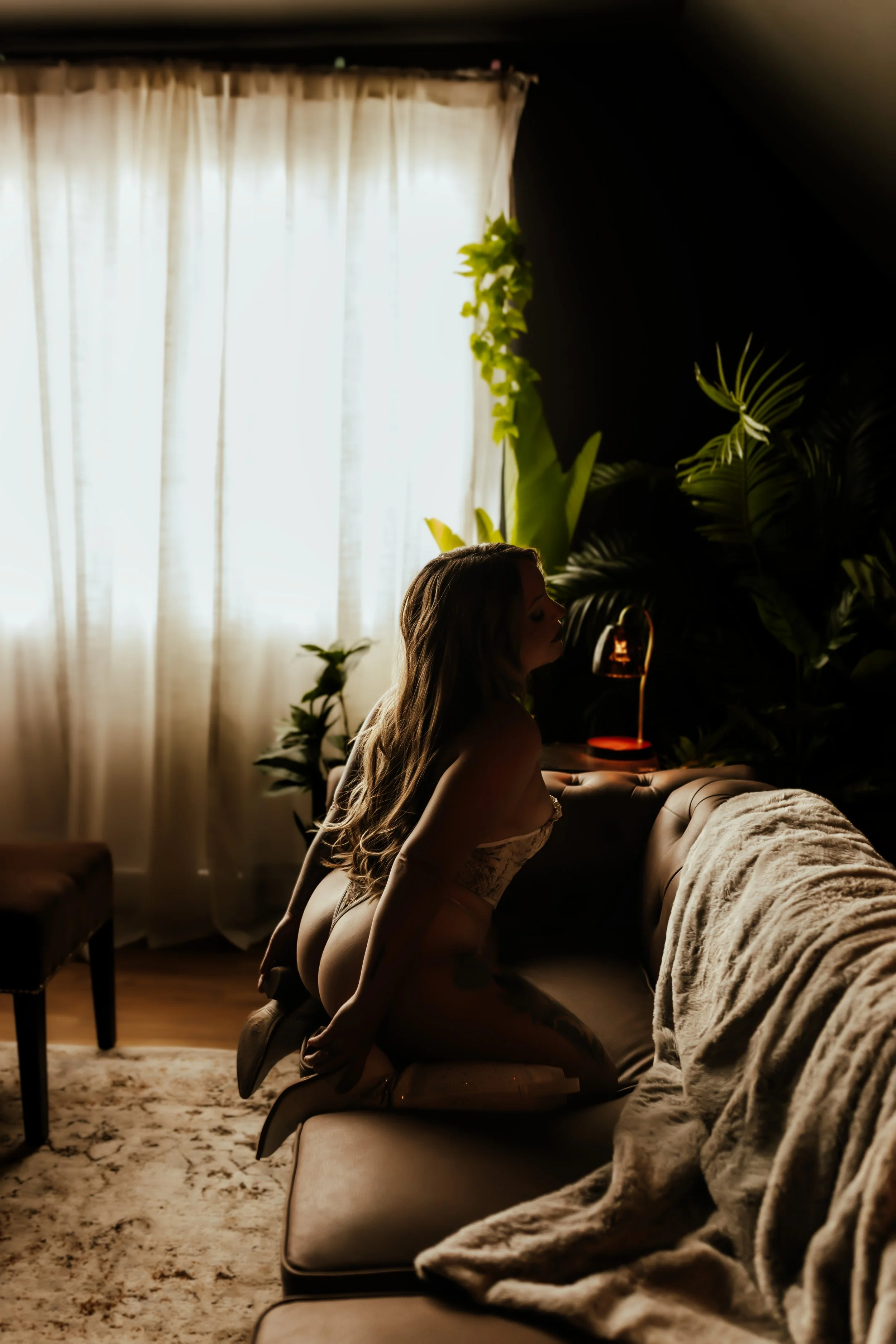 A woman with long hair kneeling on a cushion on a beige carpet in a dimly lit room, surrounded by large green plants, with a beige tufted sofa and cream curtains in the background.