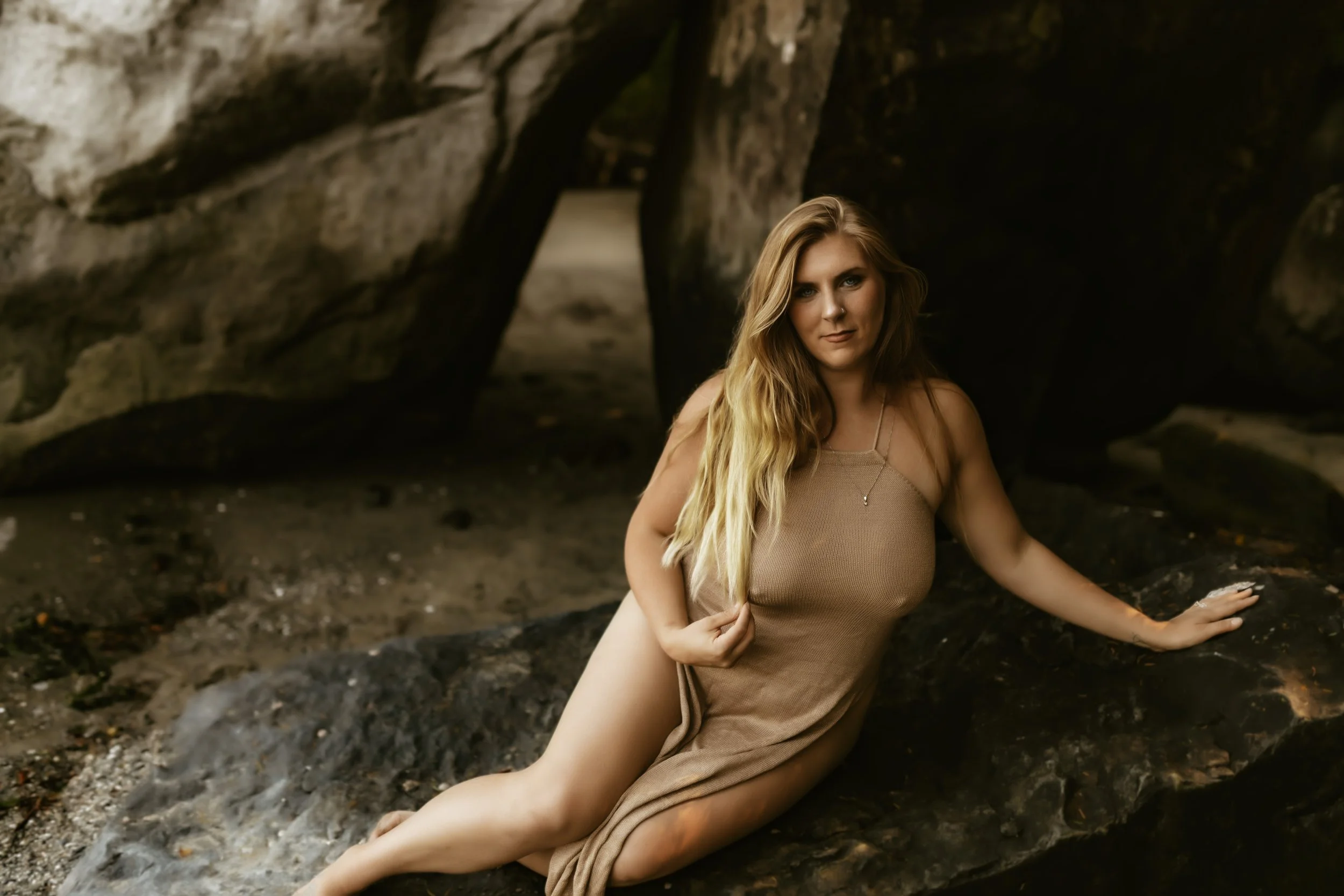 A woman with long blonde hair sitting on the ground in front of a large rocky cave opening.
