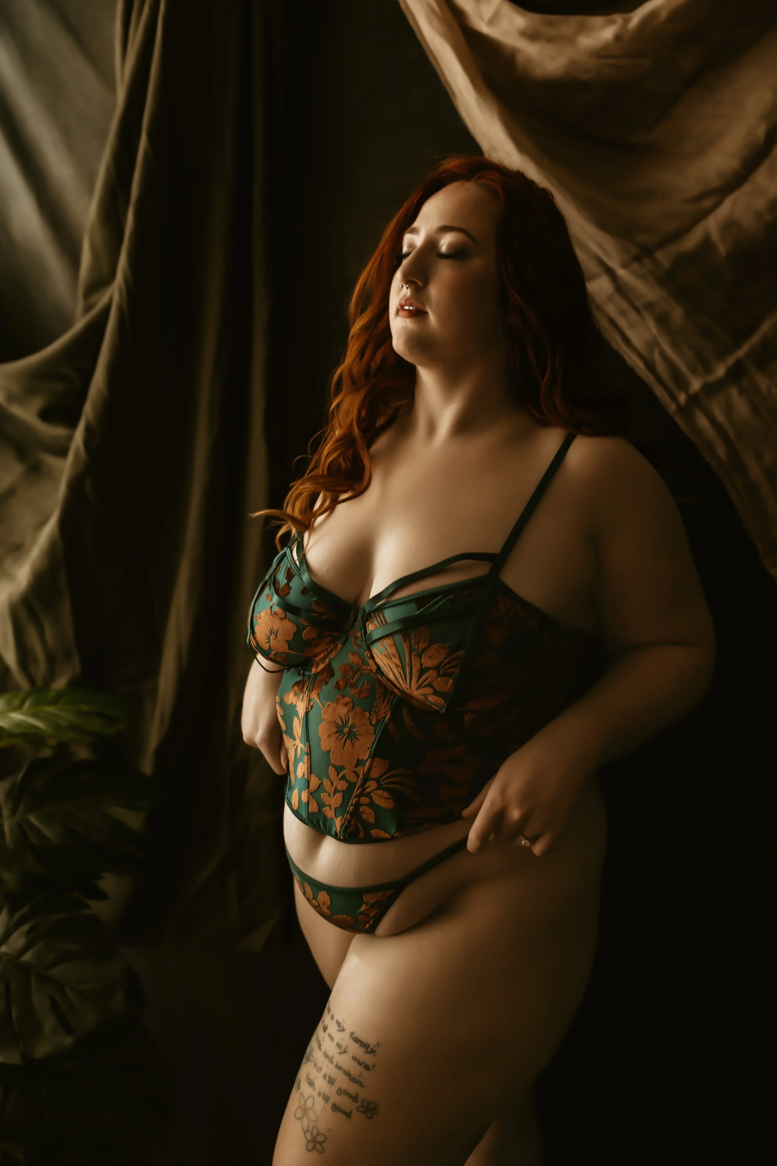 Woman with red hair and tattoos on her thigh wearing floral lingerie, standing with eyes closed in front of dark background with draped fabric and green tropical plants.
