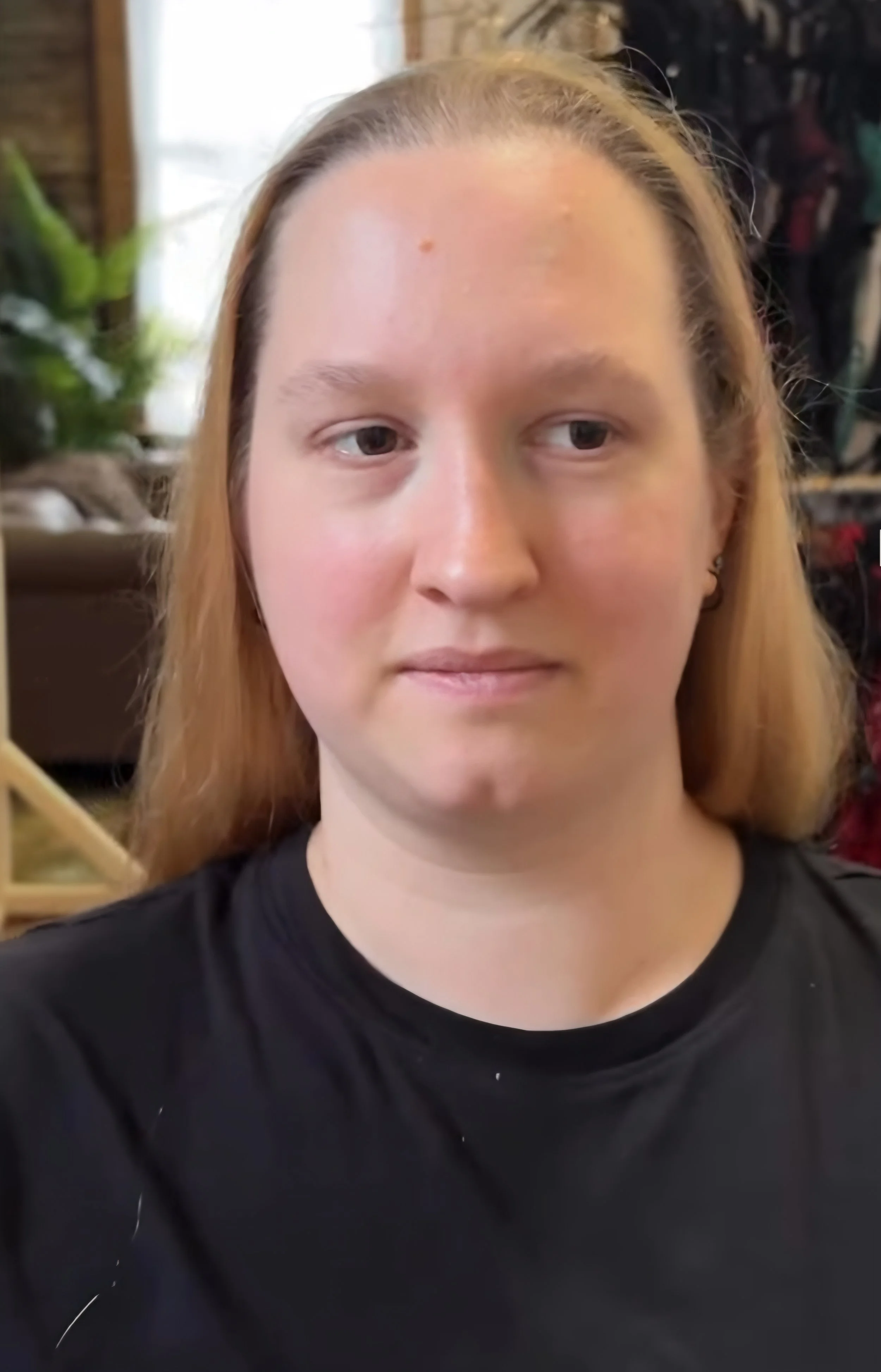 Close-up of a young woman with blonde hair, wearing a black T-shirt, standing indoors in front of a blurred background. Minimal facial expression.