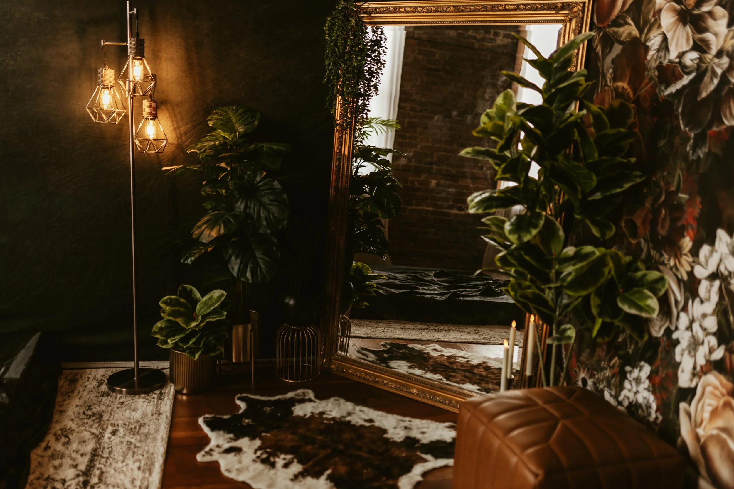 A cozy interior with a large gold-framed mirror, lush green plants, a cowhide rug, and a cowhide ottoman, illuminated by warm lighting from a modern geometric chandelier.