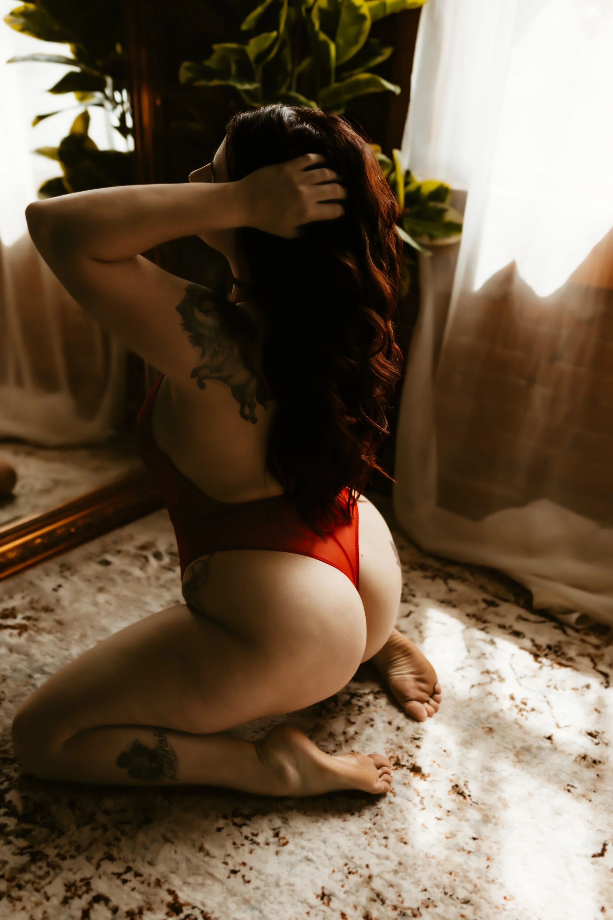 A woman with long hair and tattoos on her arm and leg, kneeling on a plush, patterned rug in front of a mirror, with her hands in her hair. She is wearing red lingerie and is positioned near a window with white curtains, in a softly lit room with pla