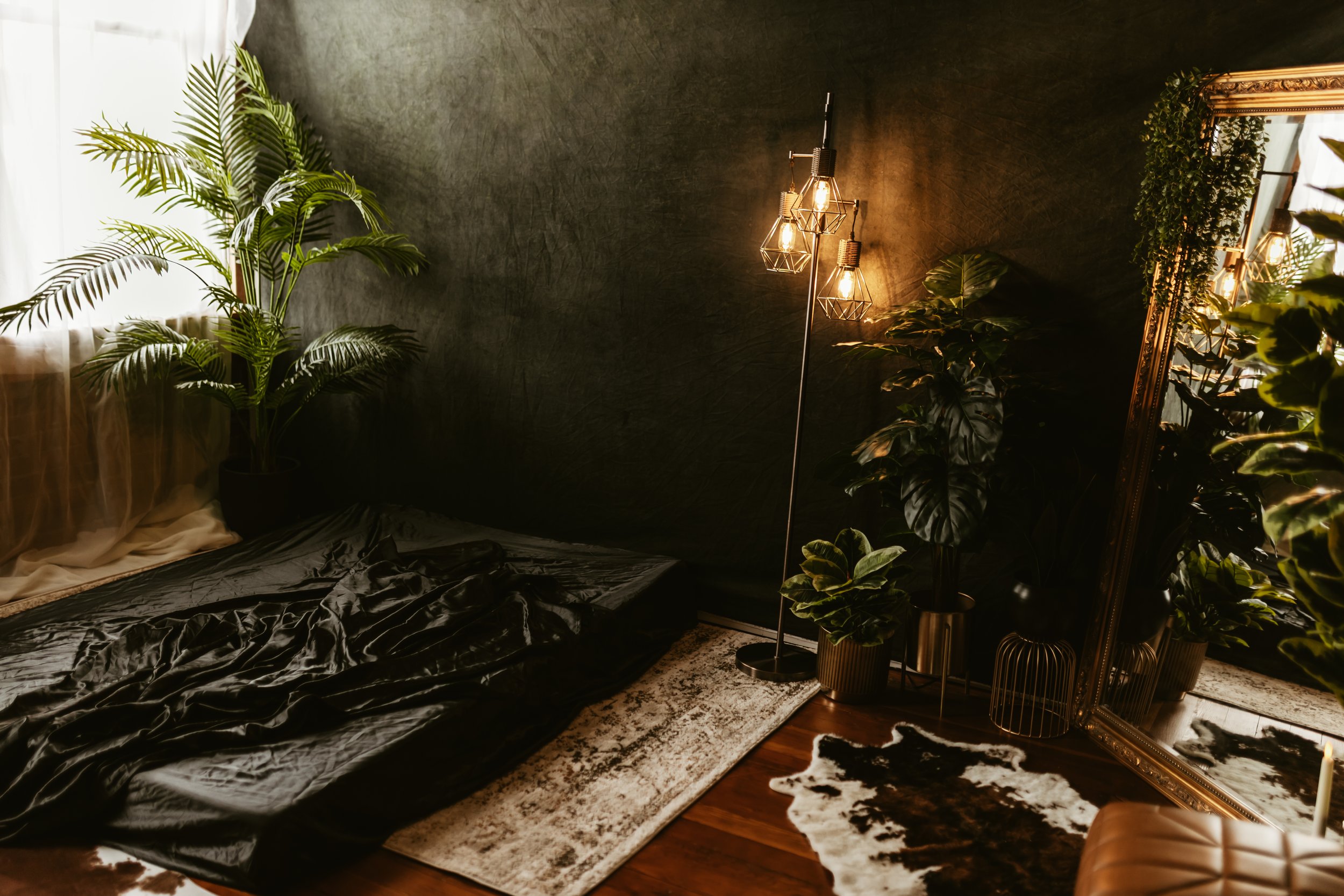 A cozy interior corner with dark walls, large green plants, a standing lamp with exposed bulbs, a large mirror, and a plush cowhide rug on hardwood floors.
