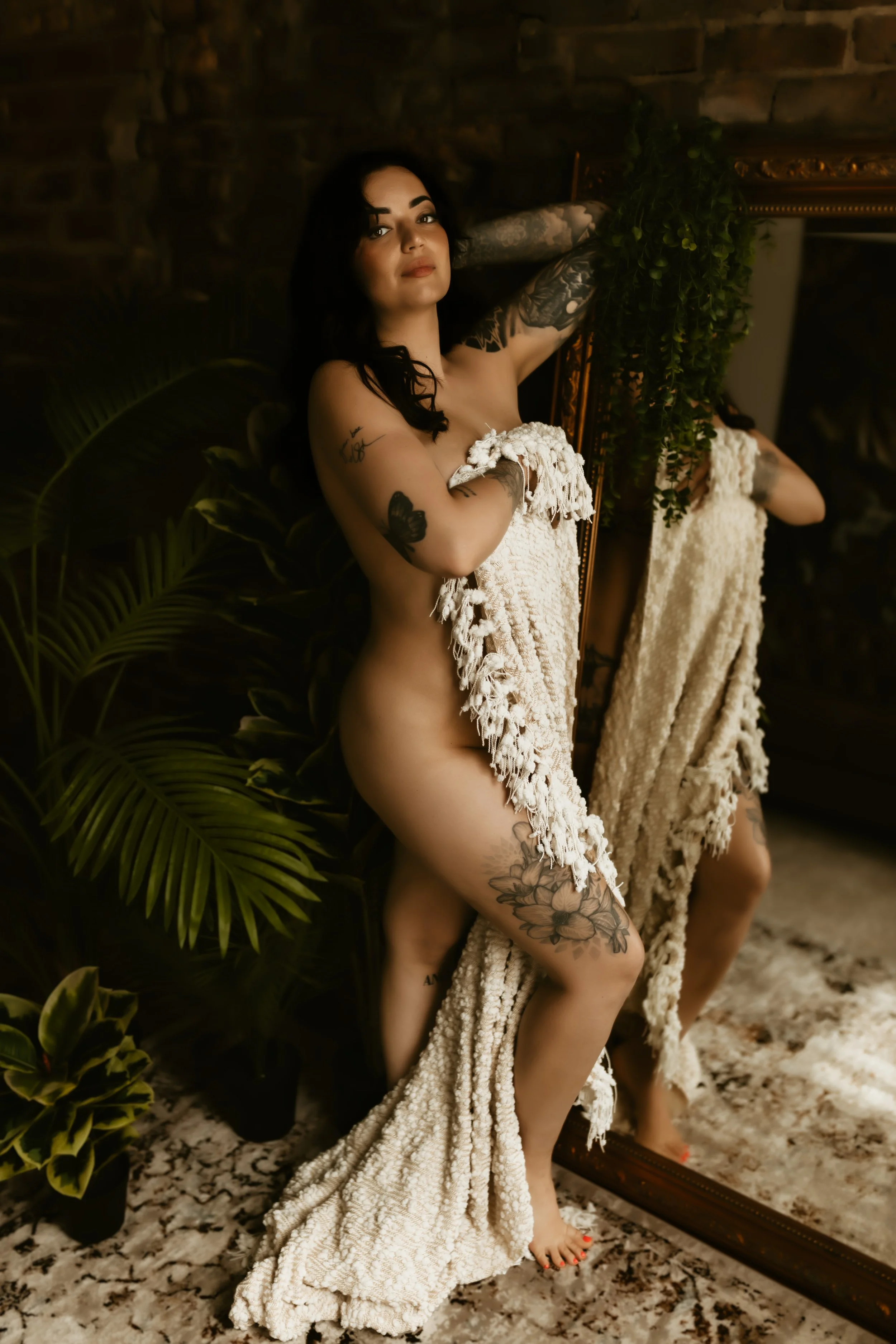 A woman with black hair and tattoos standing in front of a mirror, wrapped in a textured beige towel, in a room with a dark brick wall, greenery, and a patterned floor.
