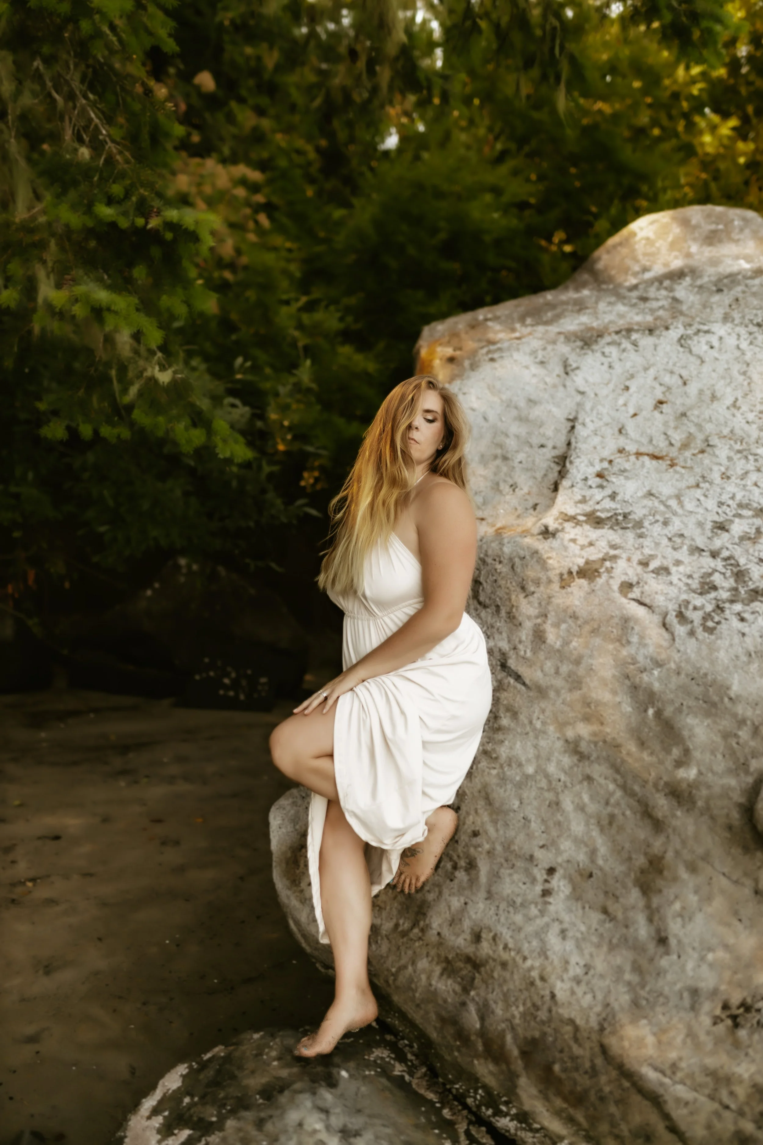 A woman with long, wavy blonde hair standing against a large rock in a natural outdoor setting with trees and greenery in the background. She is wearing a white dress and is barefoot, with one foot on the rock and the other touching the ground.