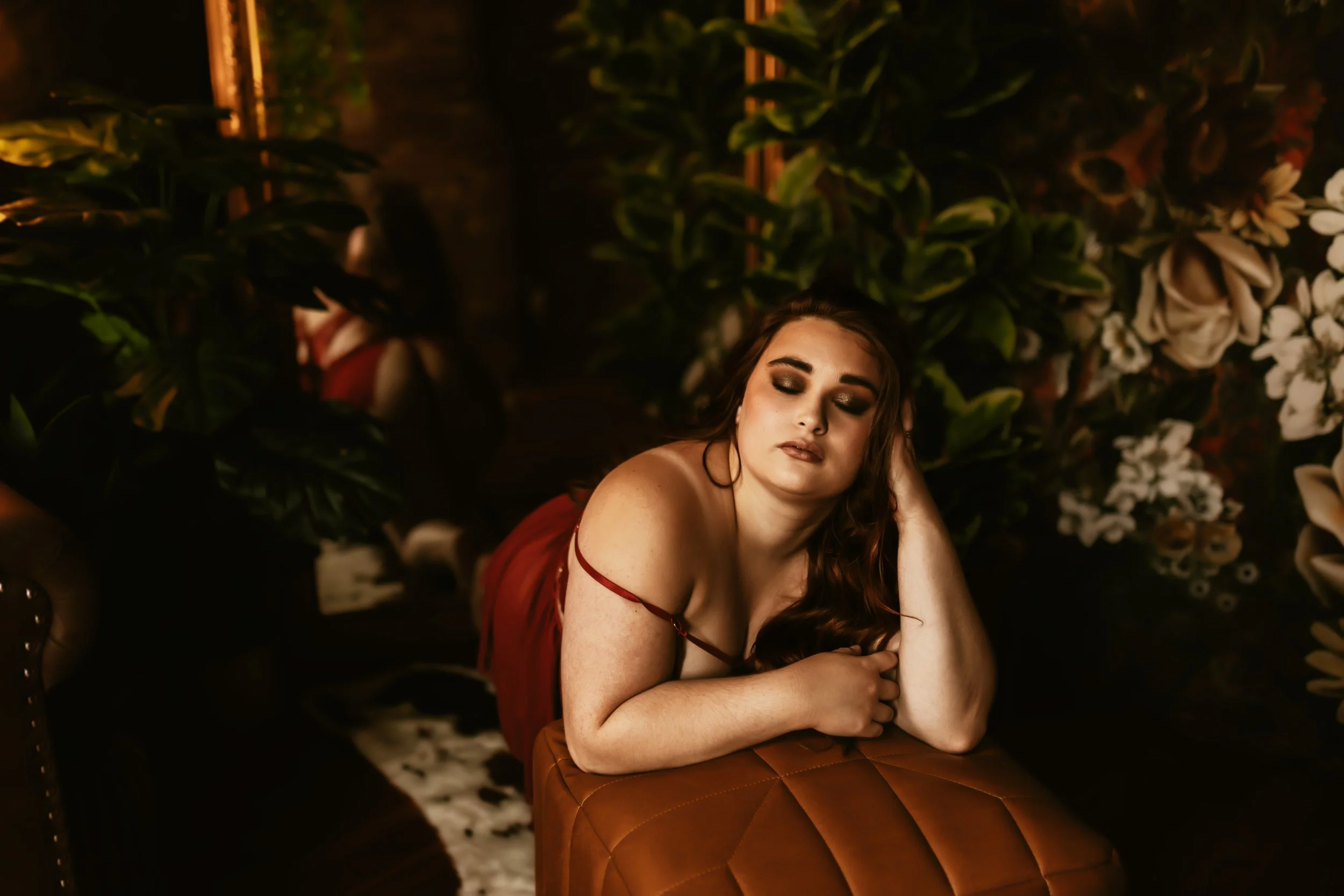 A young woman with long brown hair, closed eyes, and a relaxed expression, leaning on her arm while sitting in a dimly lit room surrounded by lush green plants and white flowers.