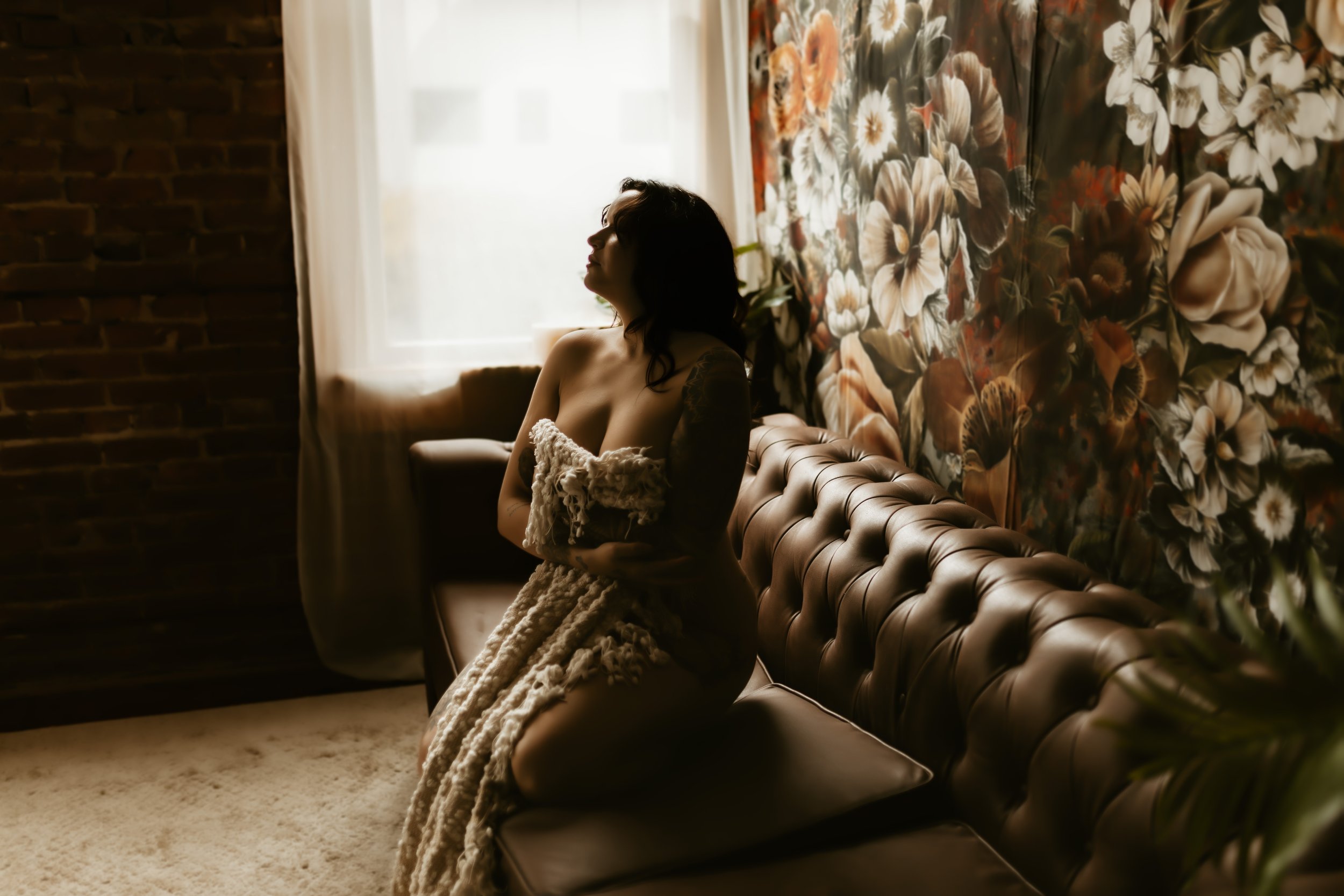 A woman with dark hair sitting on a leather tufted sofa by a window with sheer curtains, holding a blanket, next to a floral wallpapered wall in a dimly lit room.