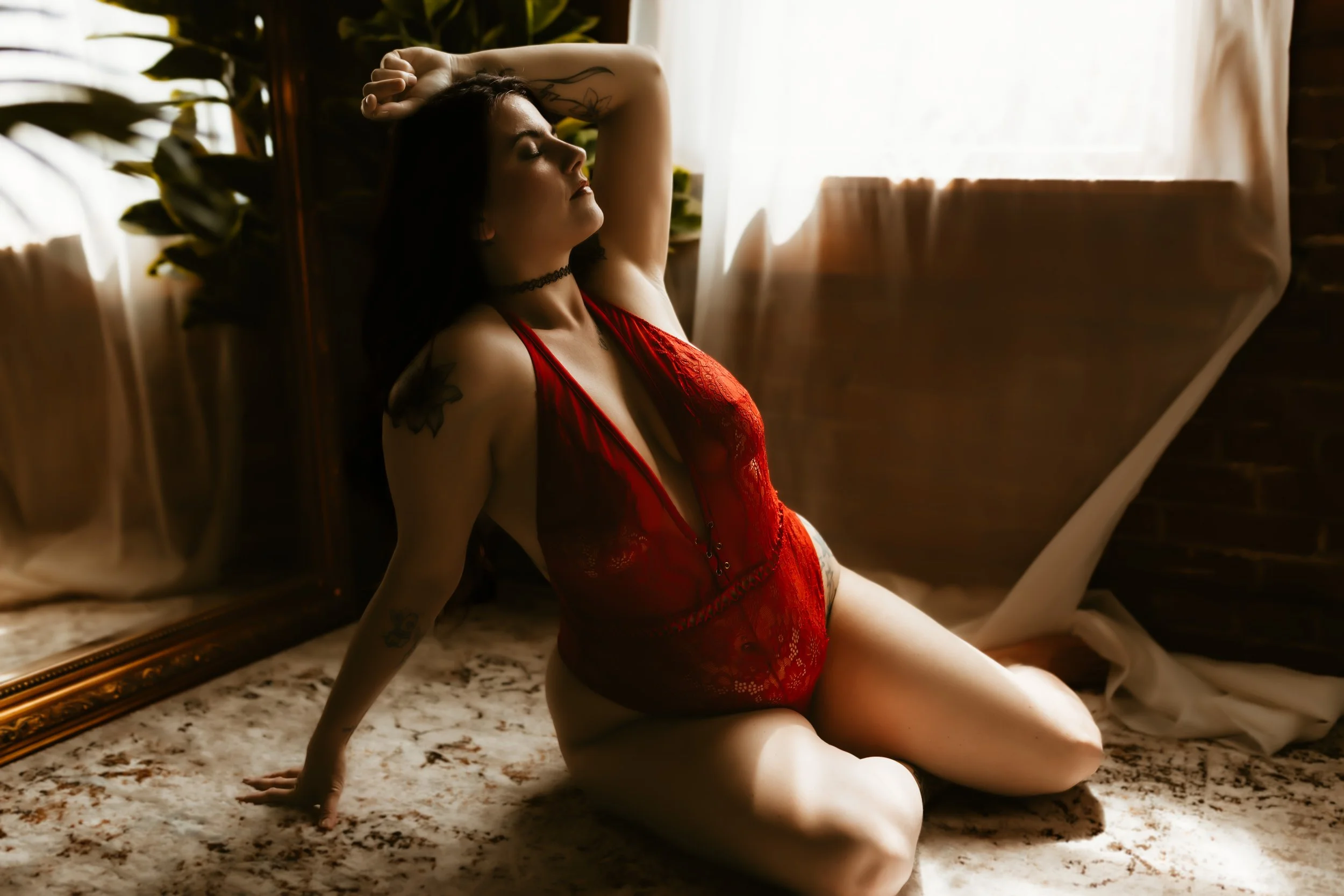 A woman in a red lace bra and underwear, sitting on the floor near a large mirror with velvet curtains and plants in the background, with her eyes closed and arm resting on her head.