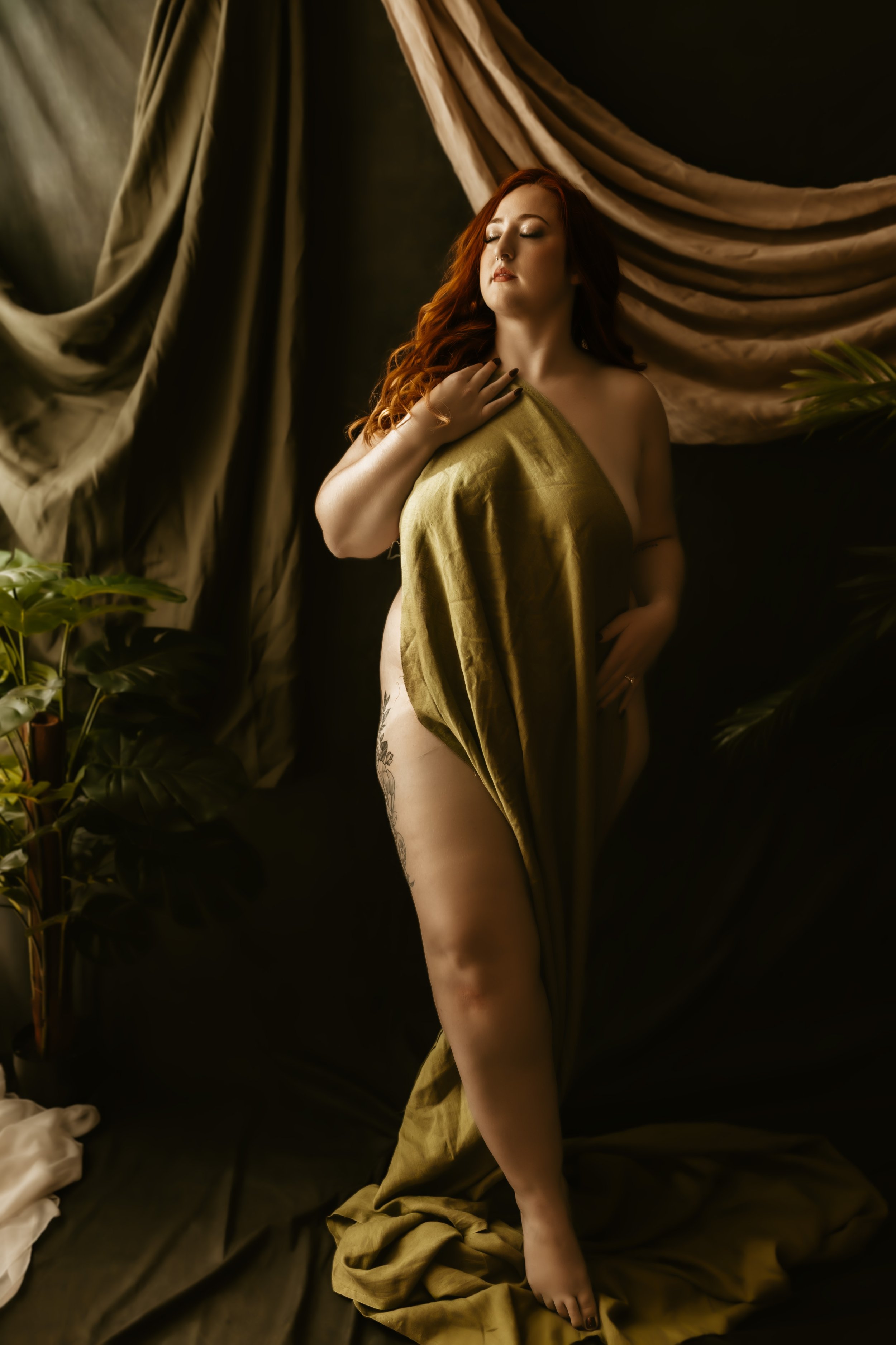 A woman with long red hair and tattoo on her leg standing on a black fabric backdrop surrounded by curtain drapes and green plants, with her eyes closed and hand resting on her chest.