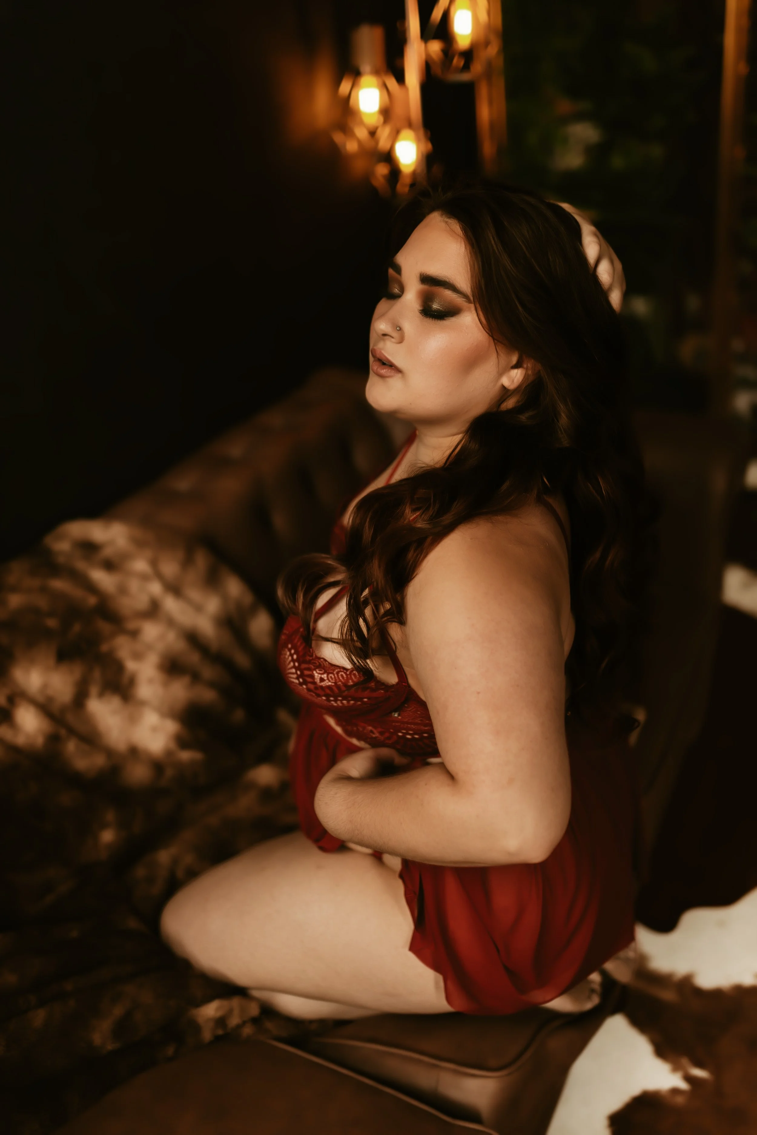A woman with dark brown hair and makeup, wearing red lingerie, sitting on a fur-covered surface with her eyes closed and hand in her hair, in a warm, dimly lit room with Edison-style light bulbs in the background.