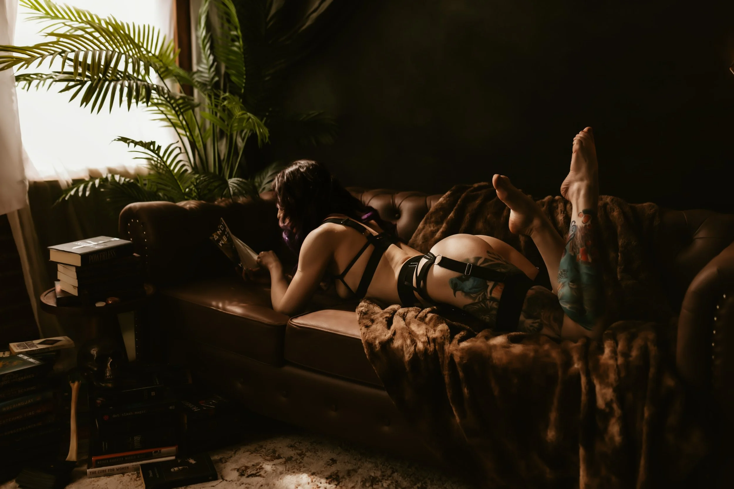 Person lying on a leather couch with a blanket, reading a book, surrounded by green plants and books on a side table.