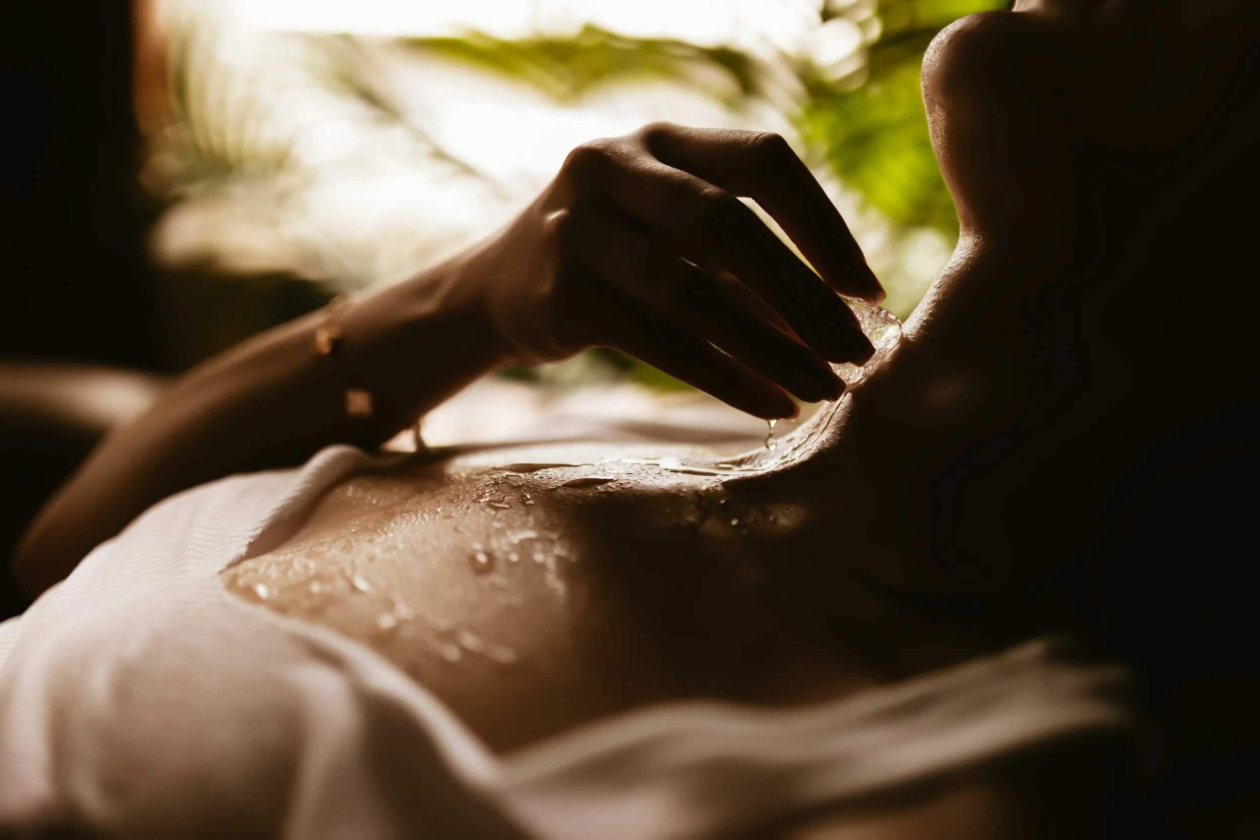 Close-up of a person receiving a back massage or skincare treatment with water or oil, with natural light and greenery in the background.
