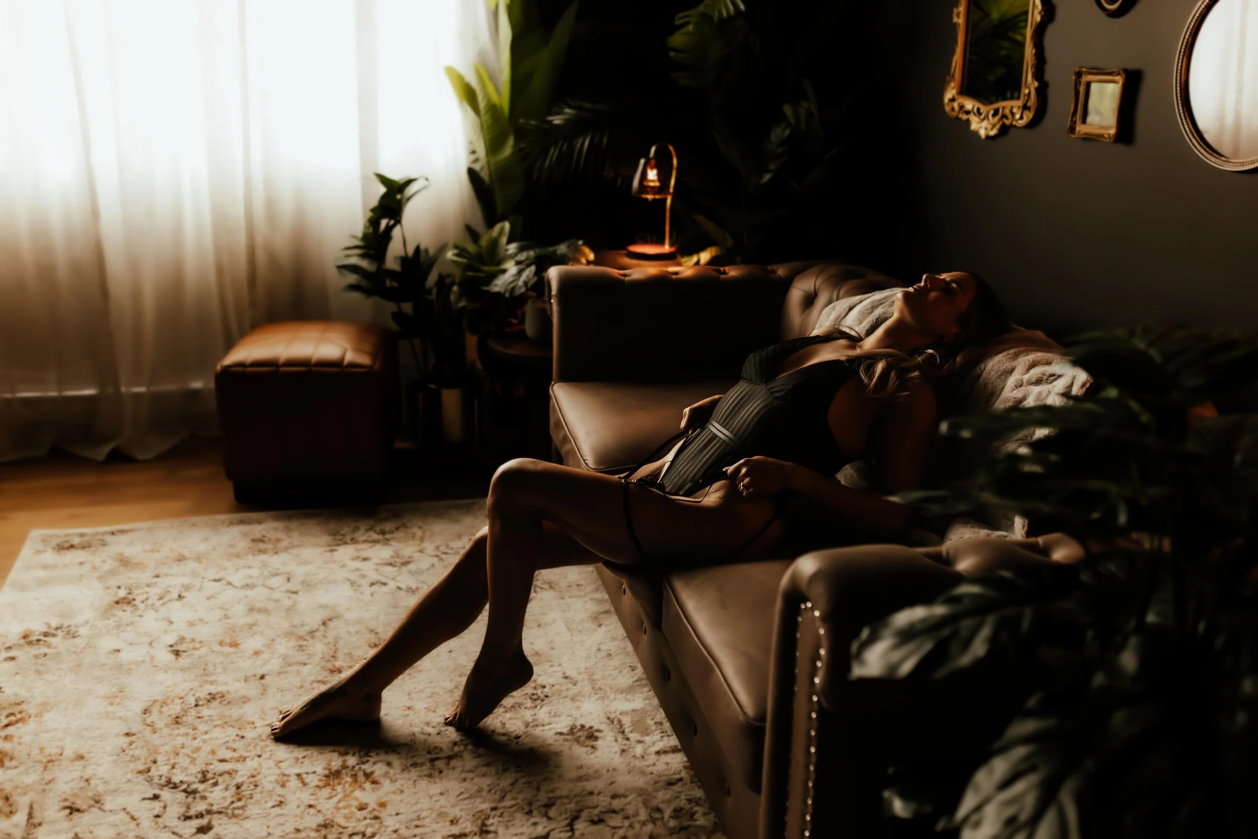 A woman wearing black lingerie sitting on a leather couch in a dimly lit room with framed mirrors on a dark wall and plants near a window with curtains.