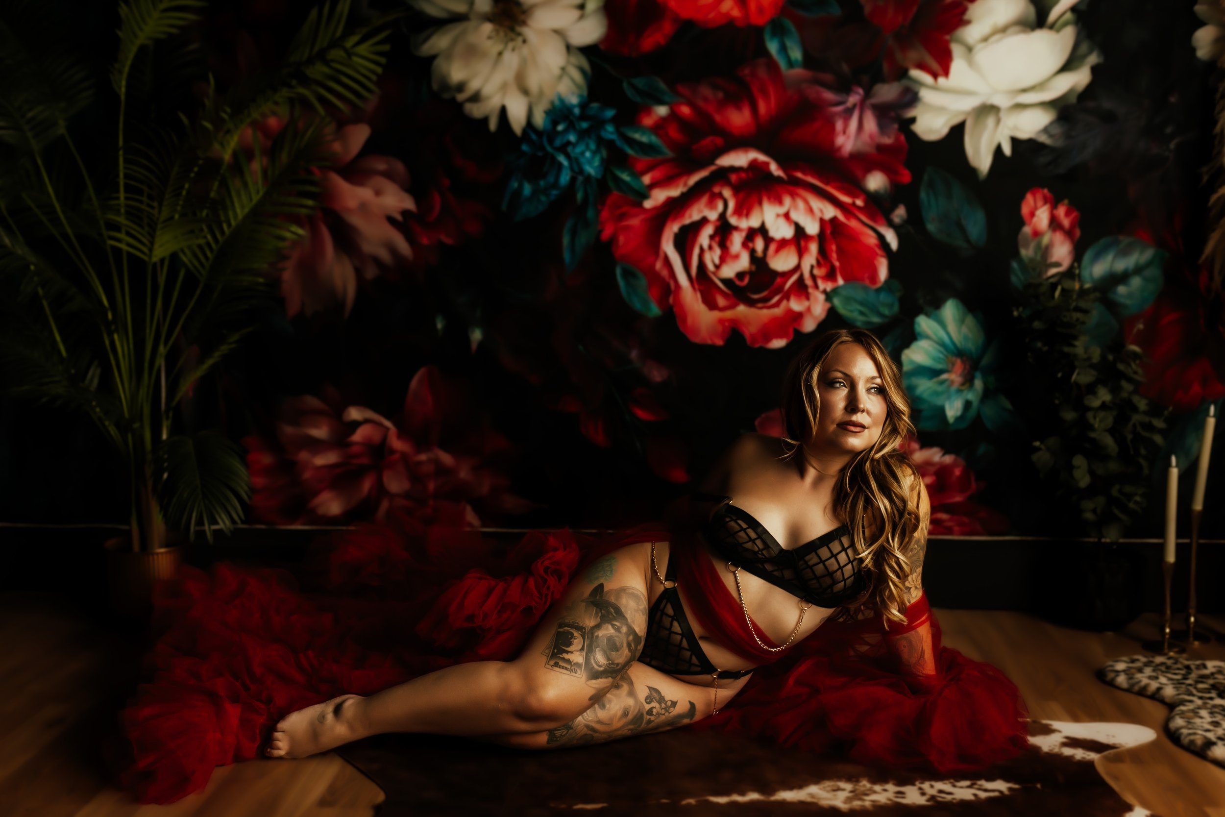A woman with tattoos and wavy hair, wearing a black and red dress, is sitting on the floor in front of a dark floral backdrop with large roses and greenery.