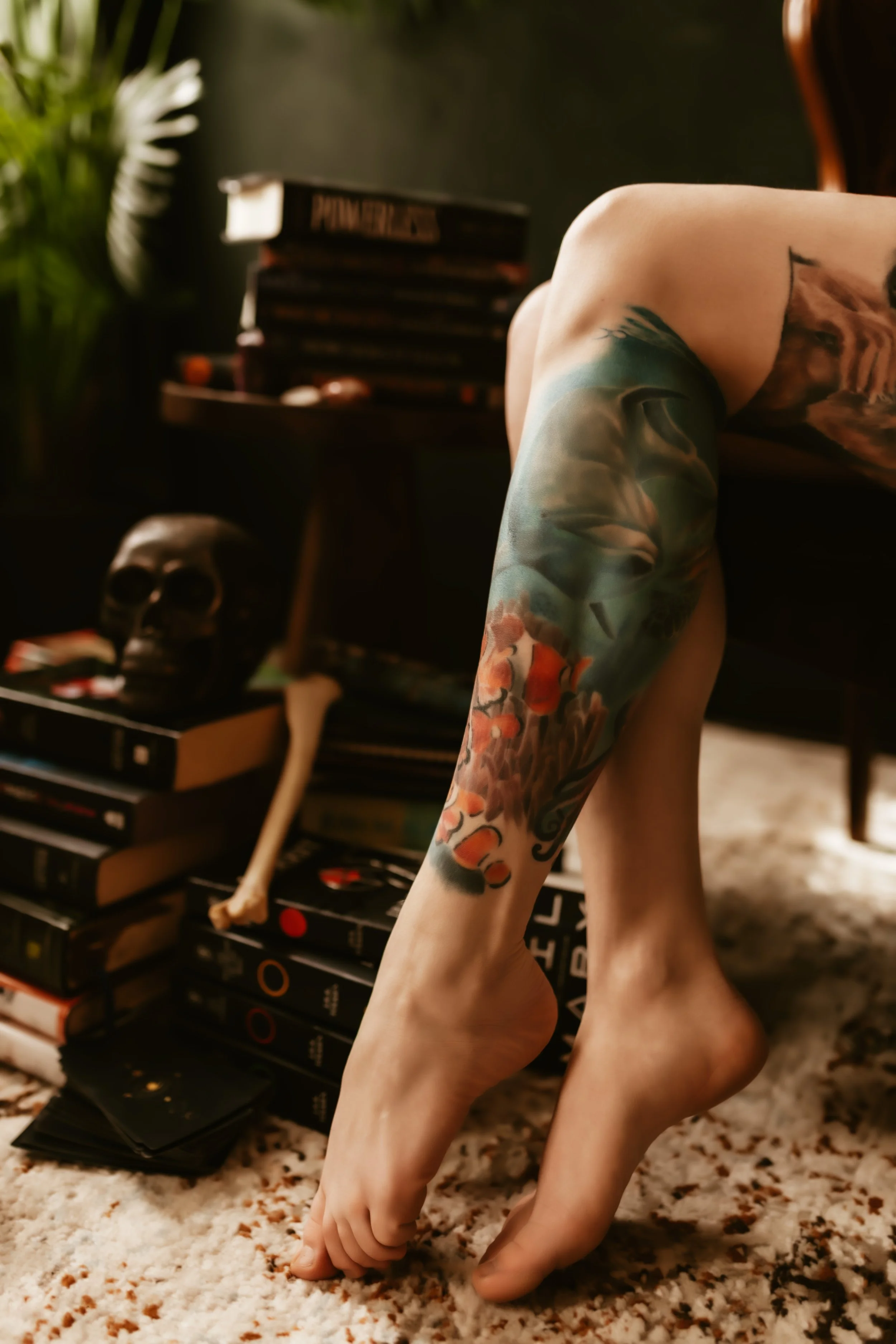 A person's foot and ankle with colorful tattoos resting on their opposite knee, in a room with a bookshelf and skull decoration in the background.