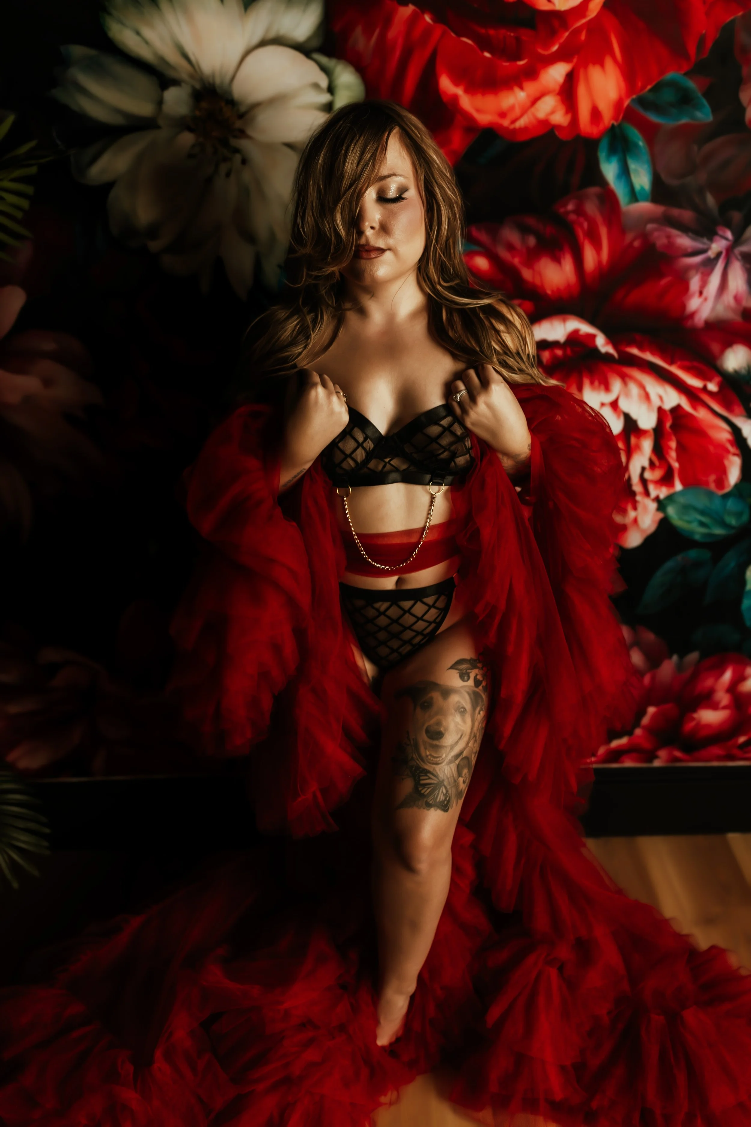A woman with long hair and tattoos on her thigh, wearing black fishnet lingerie and a red sheer robe, lying on a bed with a floral backdrop that features large white and red flowers.