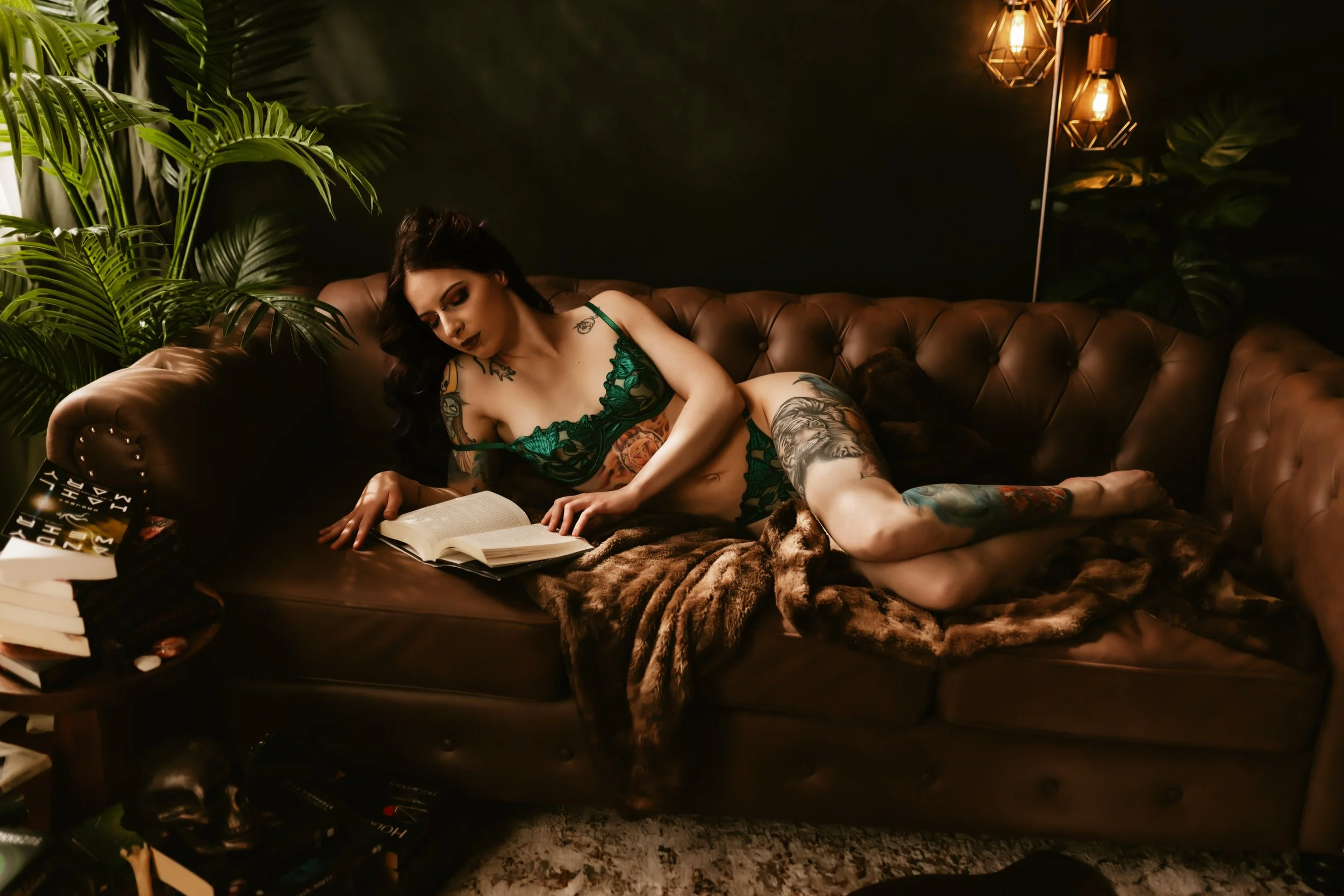 A woman with tattoos, wearing green lace lingerie, is lying on a brown leather sofa in a dimly lit room with dark walls and warm lighting fixtures. She is reading a book.