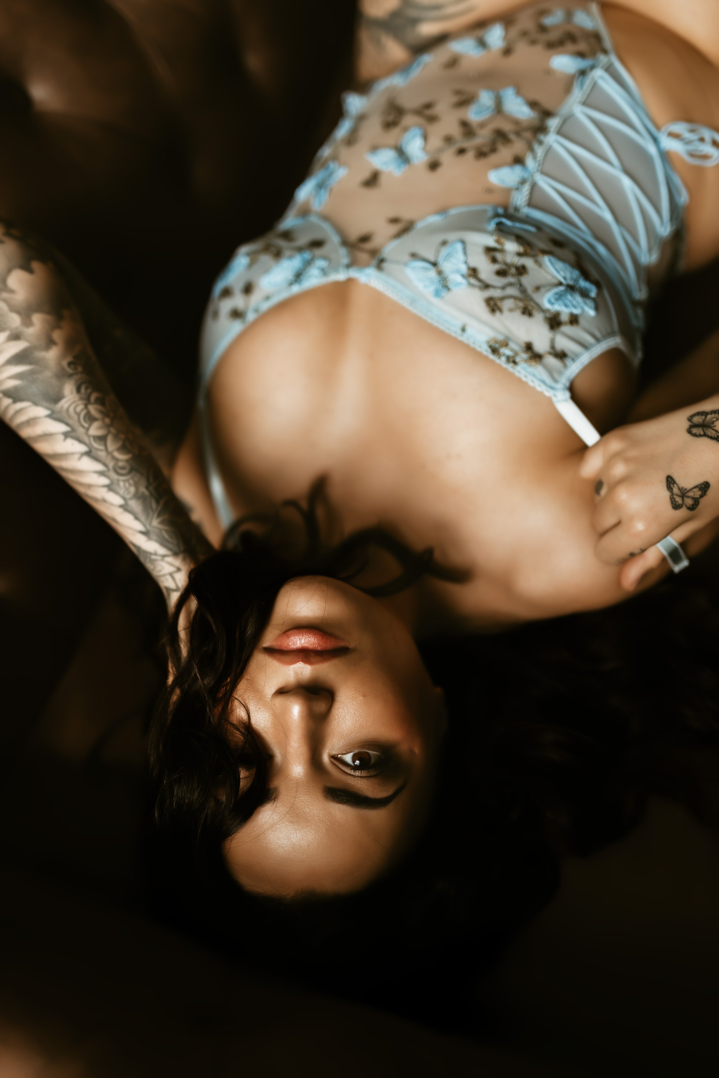 A woman with dark, curly hair lying on a dark surface, wearing lace and floral-patterned lingerie, with tattoos on her arms, looking directly at the camera.