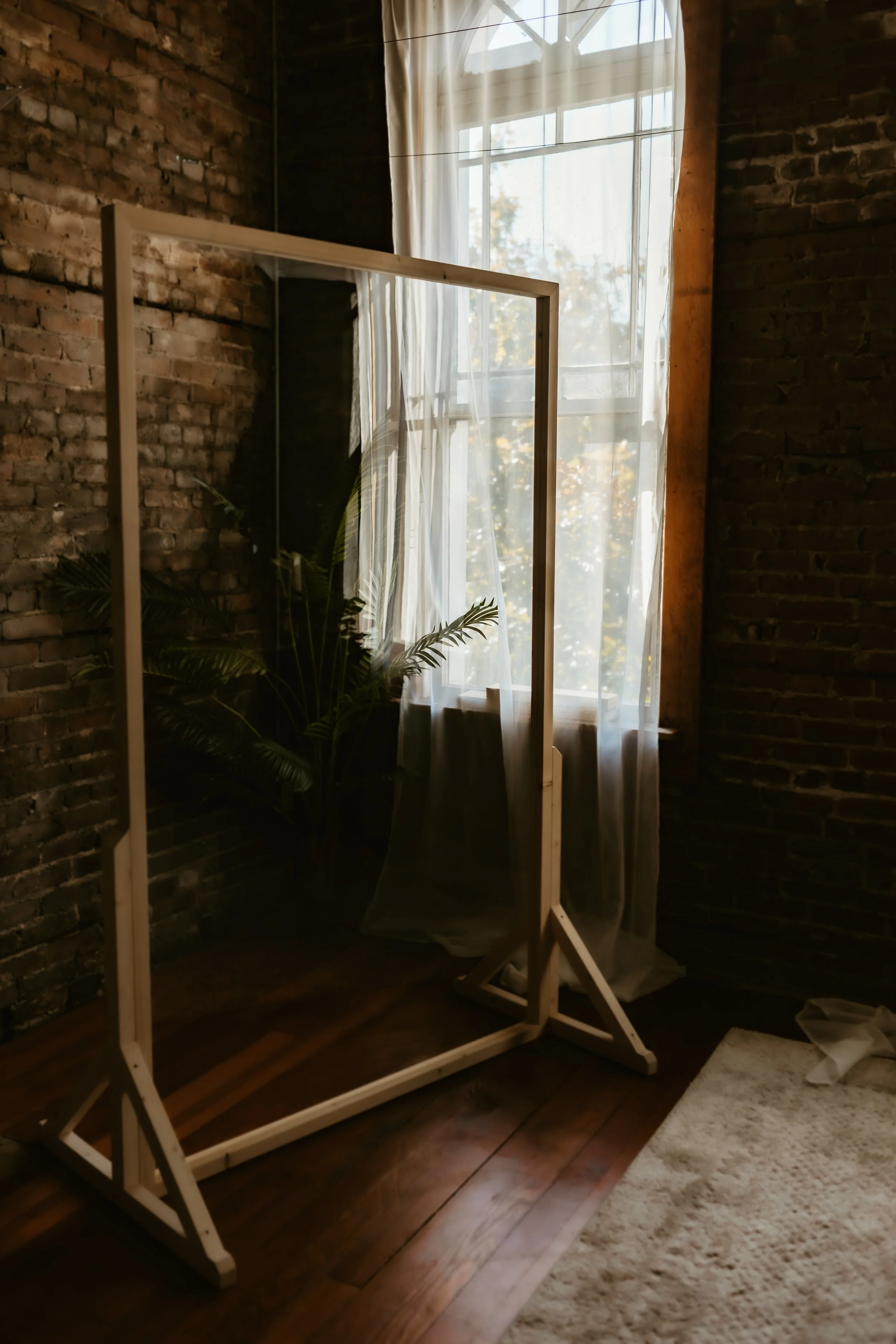 A wooden room divider standing in front of a large window with sheer white curtains, a brick wall, a potted plant, and a white rug on wooden flooring.
