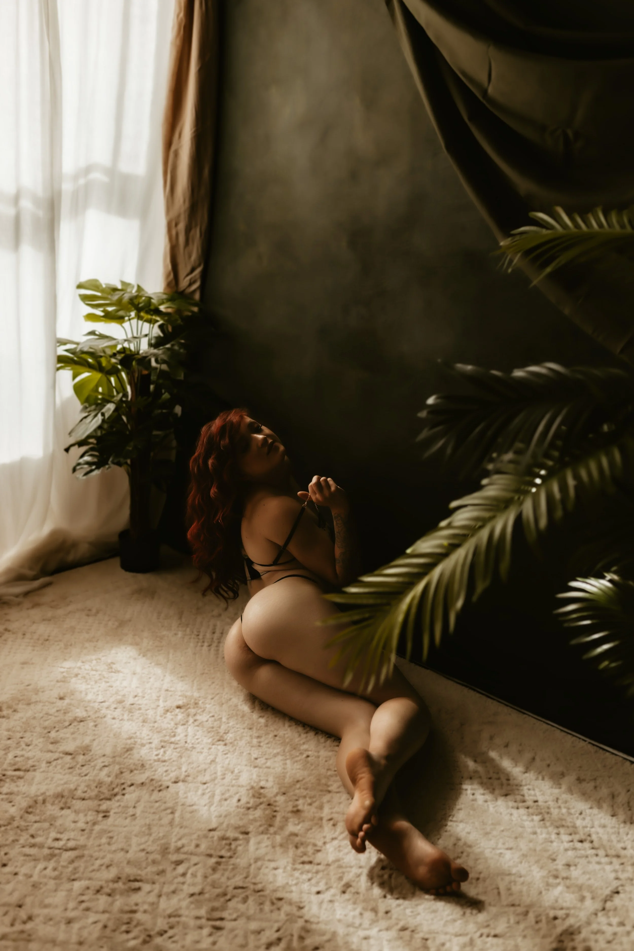 A woman with long, wavy red hair sitting on a beige carpeted floor in a room with natural light coming through a window with sheer curtains. She is surrounded by large green plants and is wearing black lingerie, with her body turned to the side and h