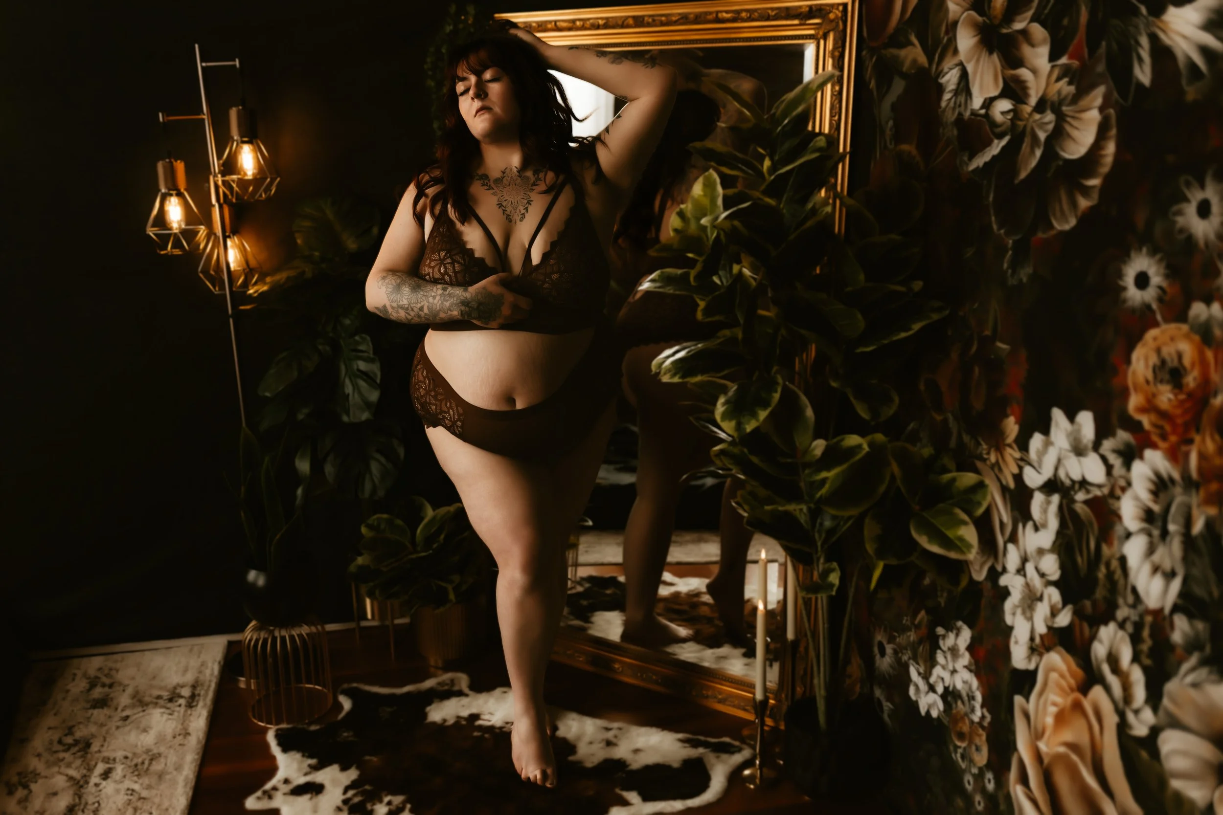 A woman wearing black lace lingerie, standing in a dimly lit room with floral wallpaper, plants, a gold framed mirror, and modern wall lamps.