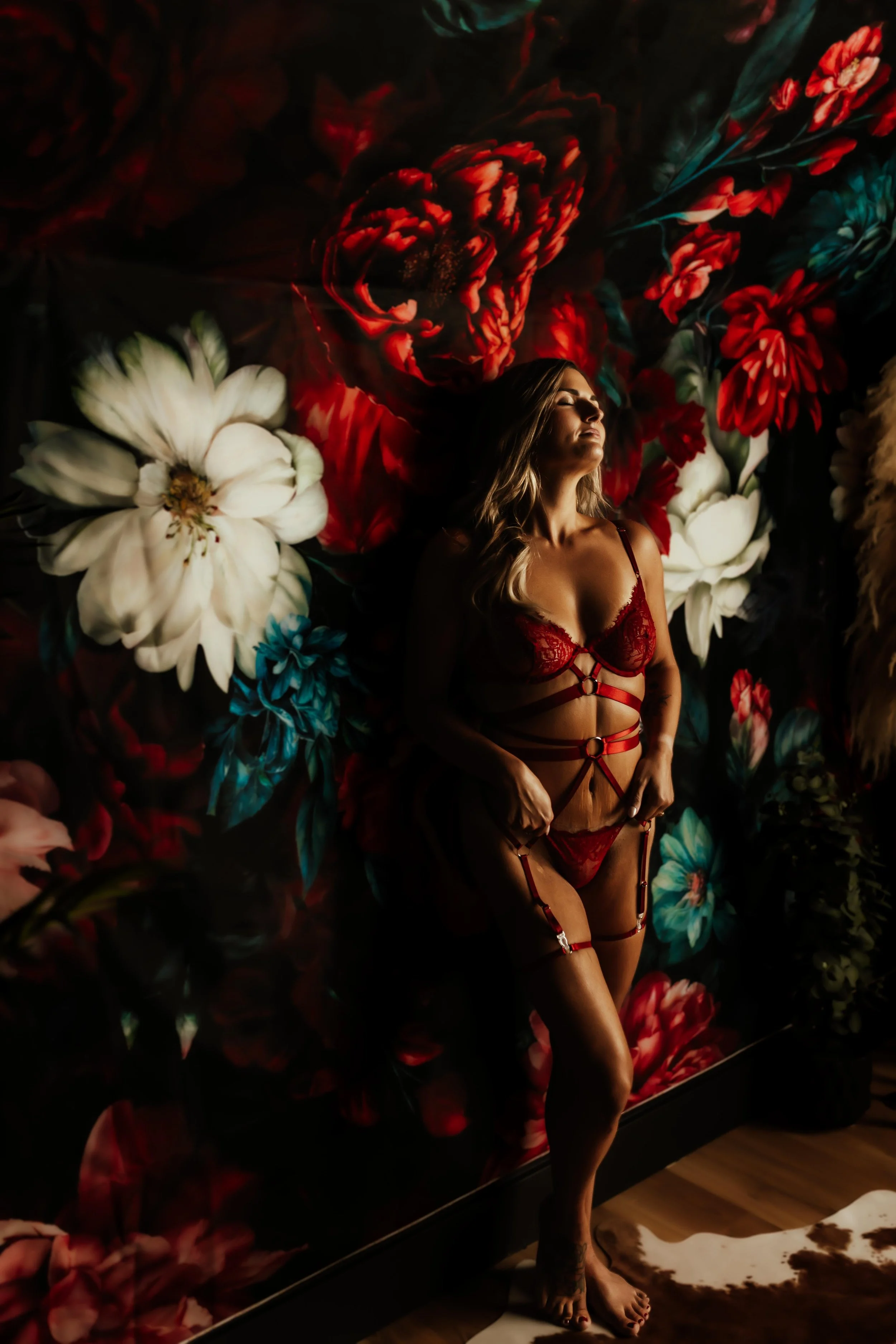 A woman in red lingerie with garter straps posing in front of a floral backdrop with white, red, and blue flowers.