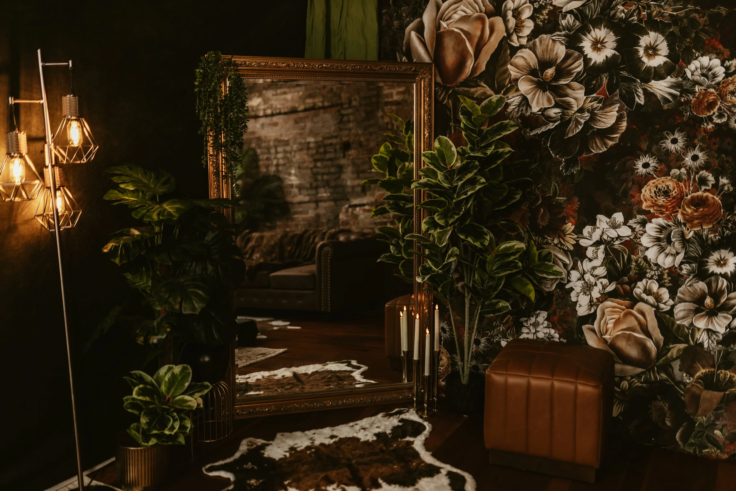 A dimly lit interior space with a floral wallpaper, large mirror, and green houseplants. There are also warm light fixtures, a cowhide rug, and a leather bench.