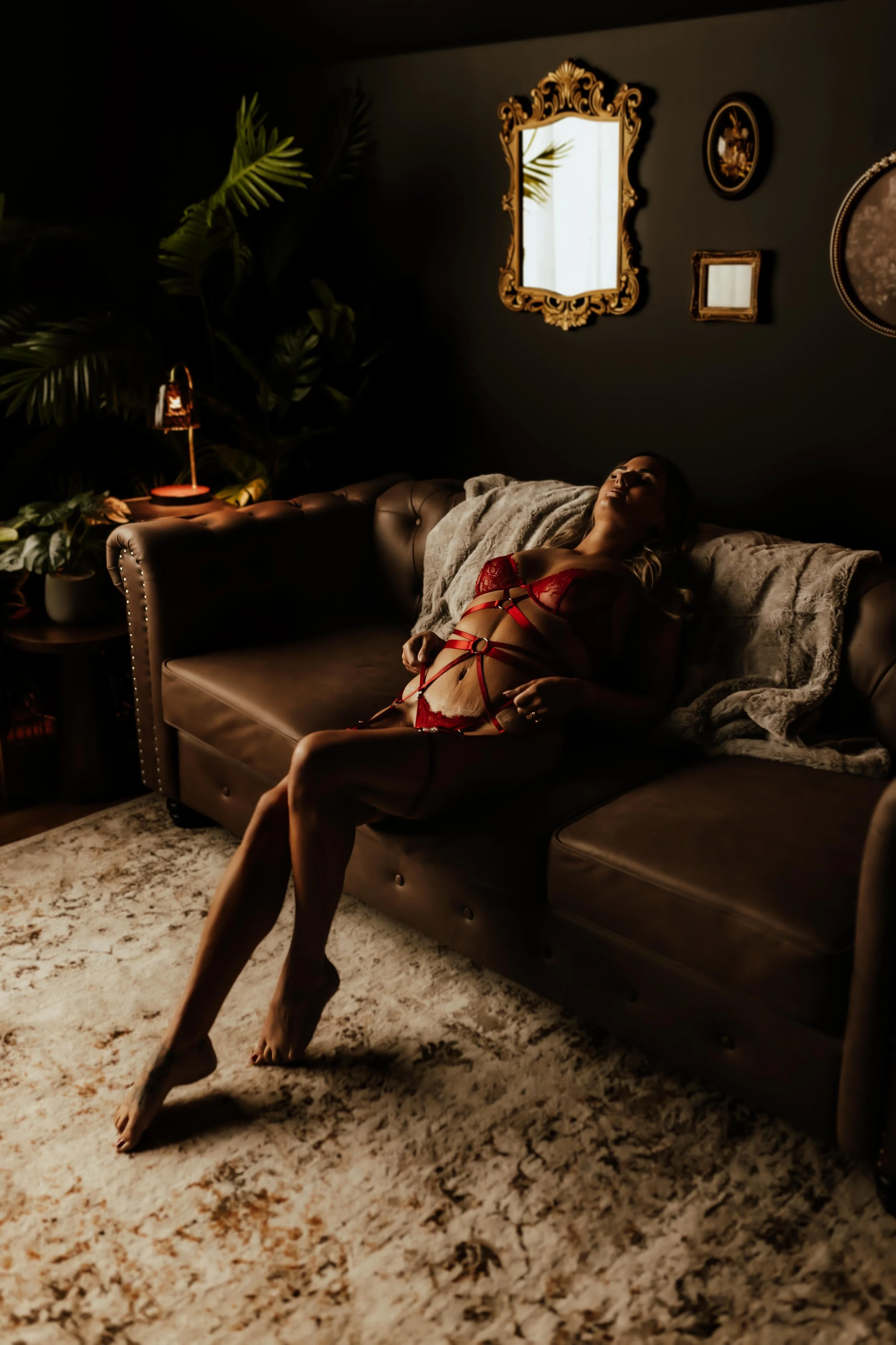 A woman with dark curly hair and tattoos on her legs sitting on a leather sofa reading a book in a dimly lit room with large green plants, a dark wall, and warm lighting.